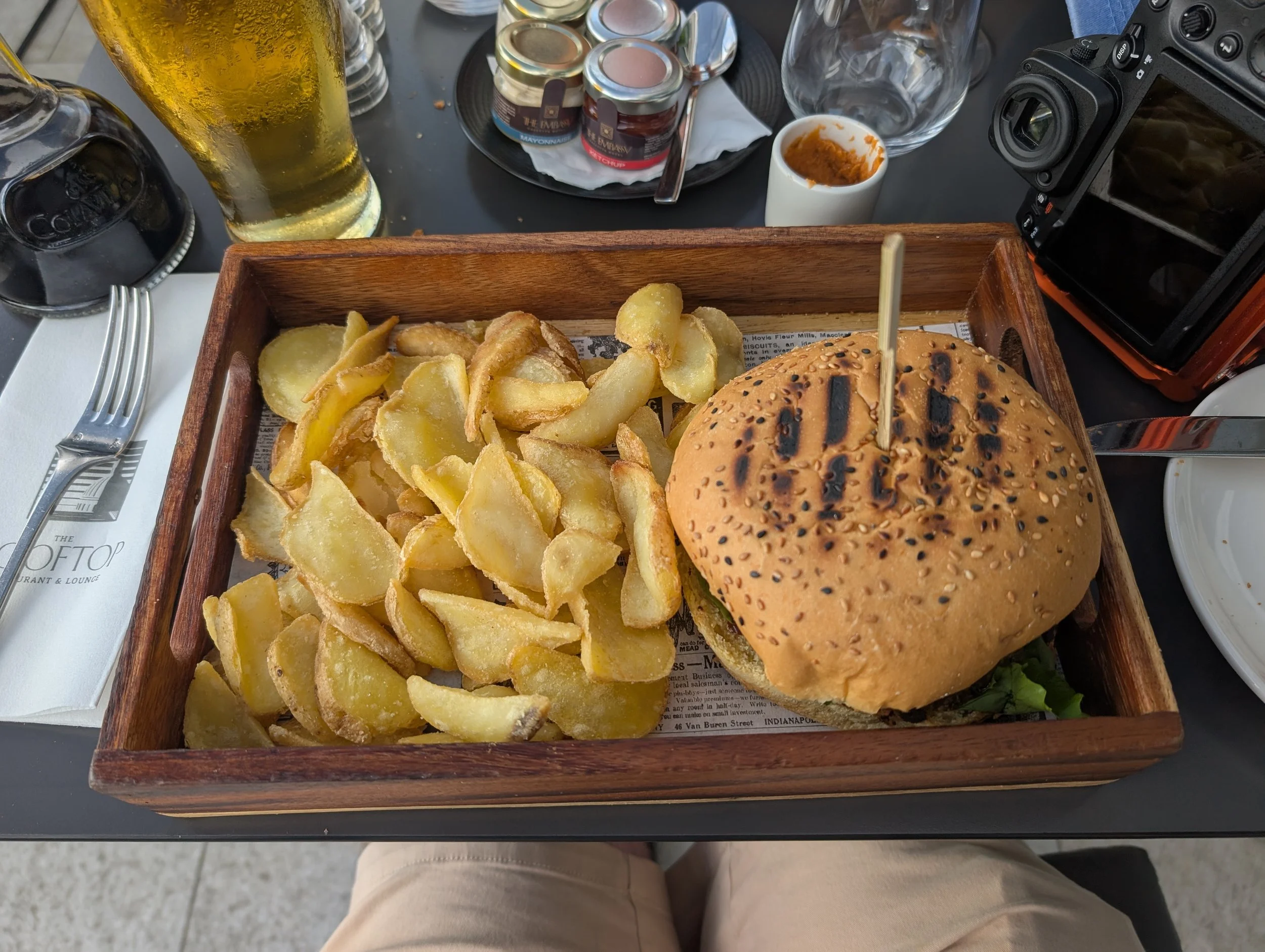 A wooden tray holding a burger with a sesame seed bun, lettuce, and a visible patty, along with a large serving of potato chips. The tray is on a table with condiments, a glass of beer, and a camera nearby.