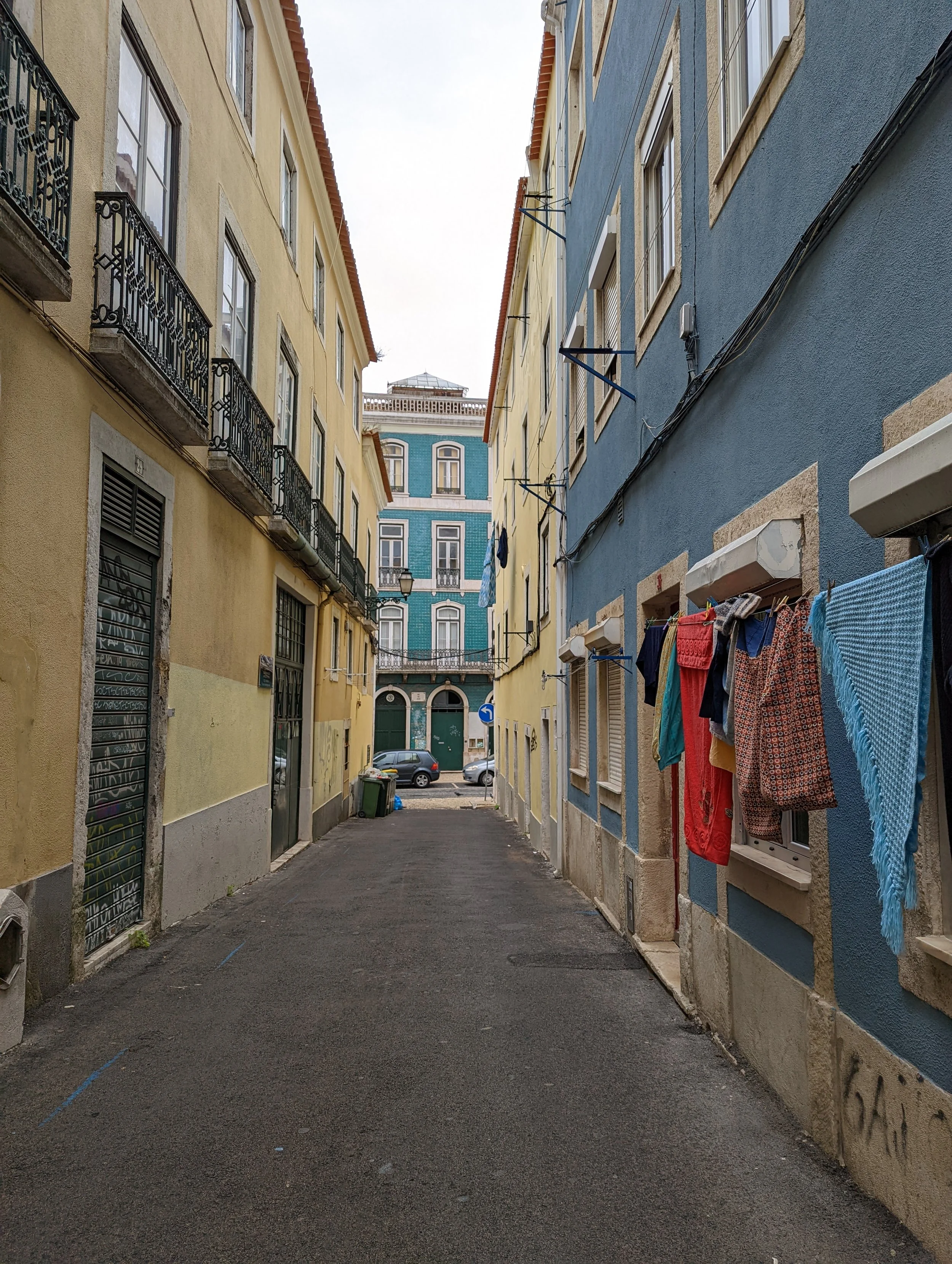 Colorful narrow street with yellow and blue buildings on either side, laundry hanging outside windows, parked cars at the end, and a 'one-way' traffic sign.