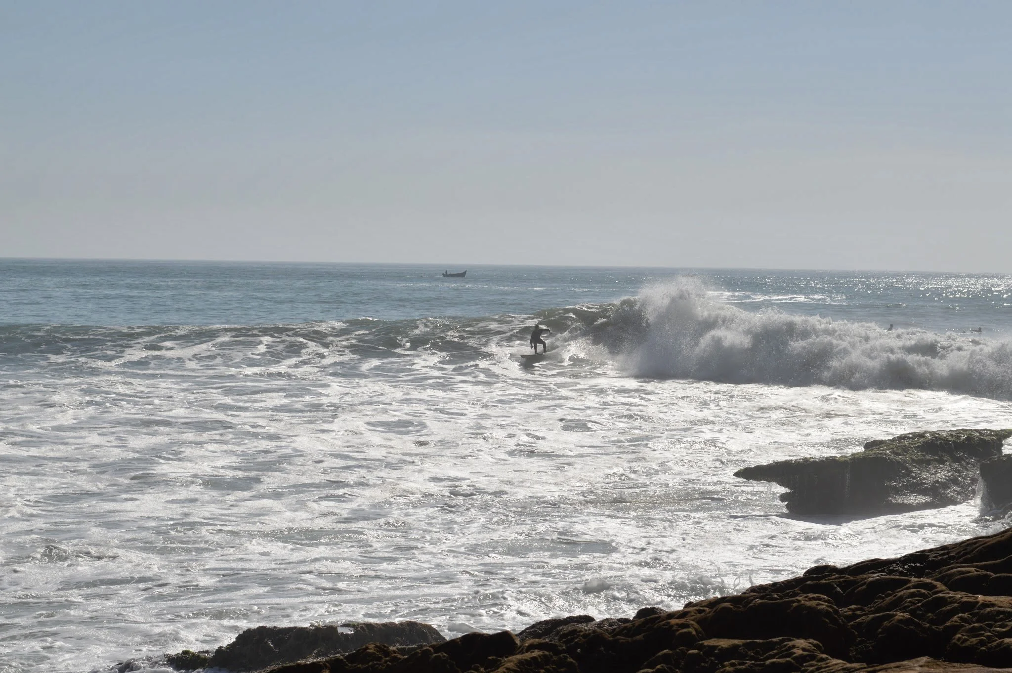 A surfer riding a wave near a rocky shoreline with a boat visible in the distance on the ocean.