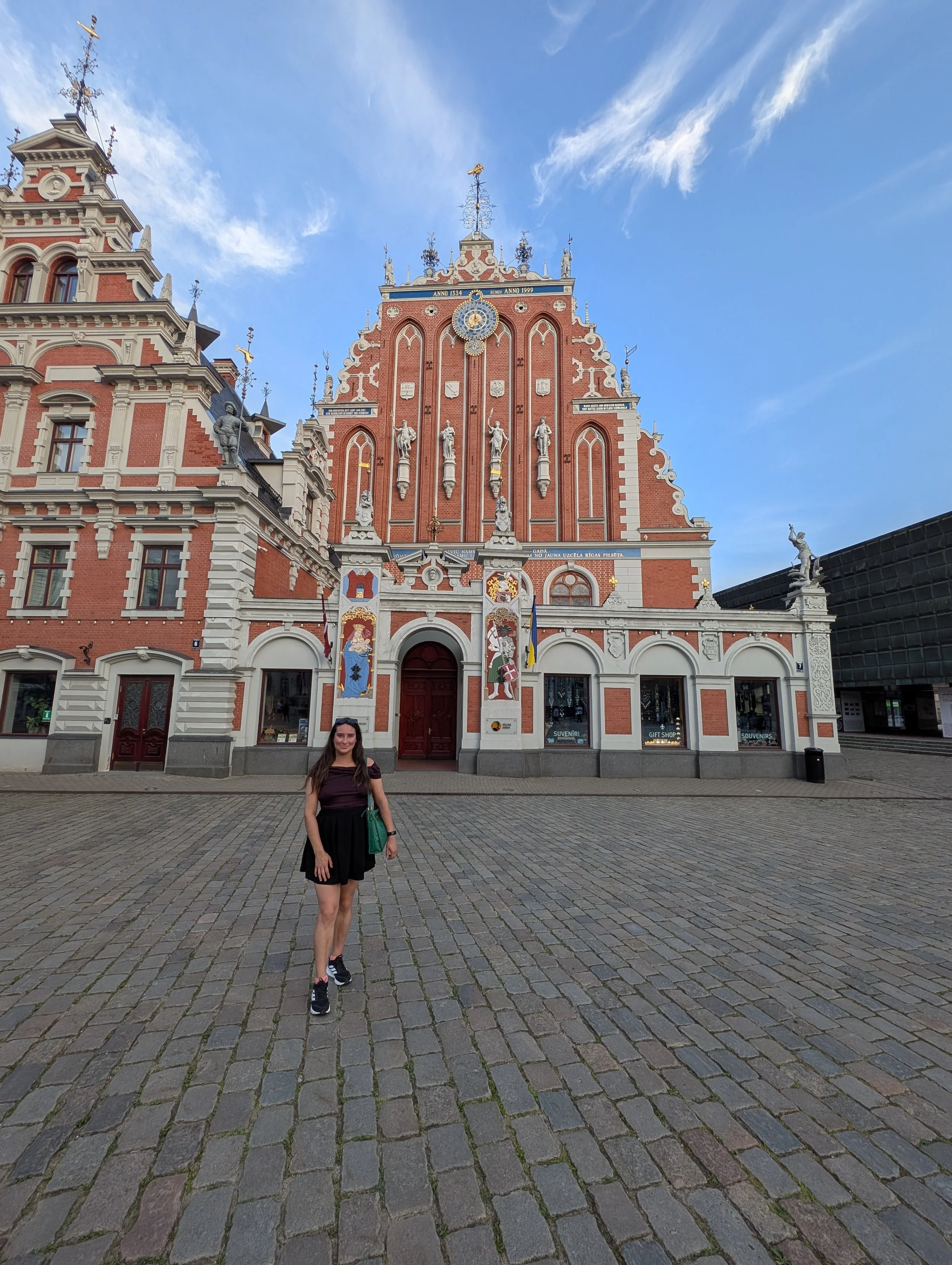 A woman with long hair wearing a black top and black skirt standing on cobblestone pavement in front of a historic building with red brick walls, sculptures, flags, and ornate architectural details, under a blue sky with some clouds.