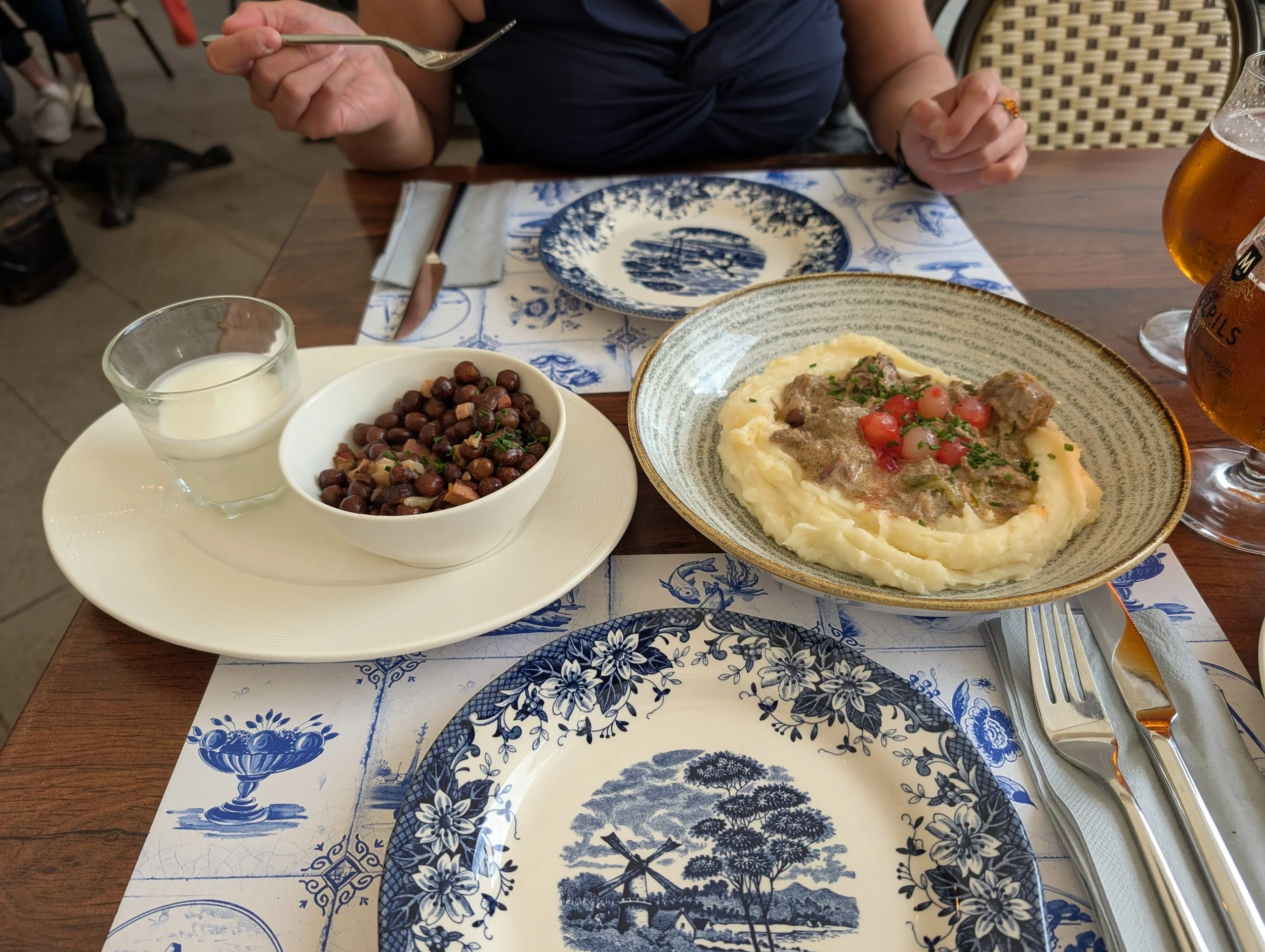 A person sitting at a table enjoying a meal with a bowl of mashed potatoes topped with beef stew and cherry tomatoes, a bowl of cooked chickpeas, a glass of a clear beverage, and a glass of beer, with blue and white patterned dinnerware and a floral 