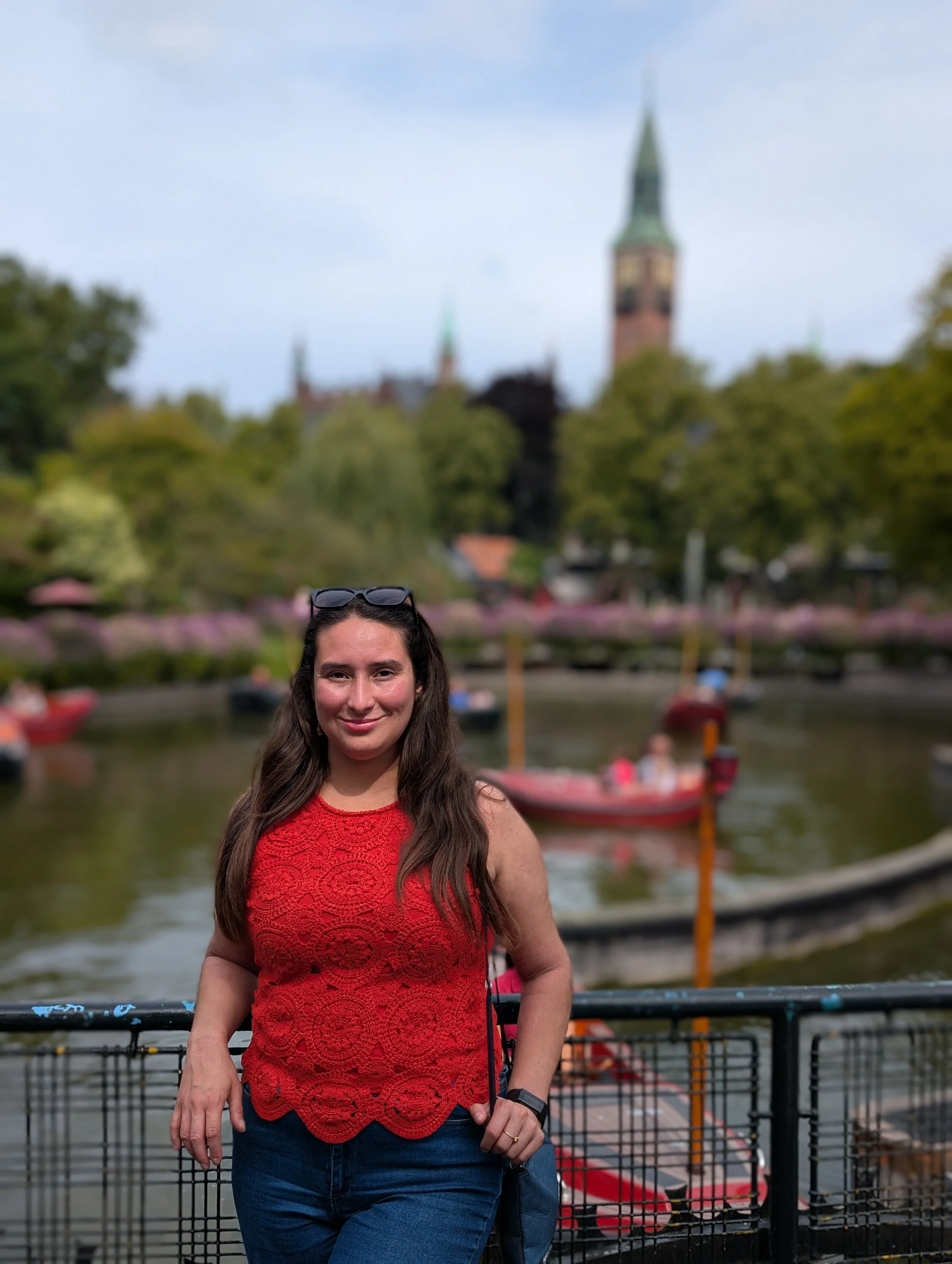 A woman with long brown hair, wearing a red sleeveless lace top and jeans, standing in front of a boat at a dock with a blurred background of a river and a cityscape, including a tall clock tower.