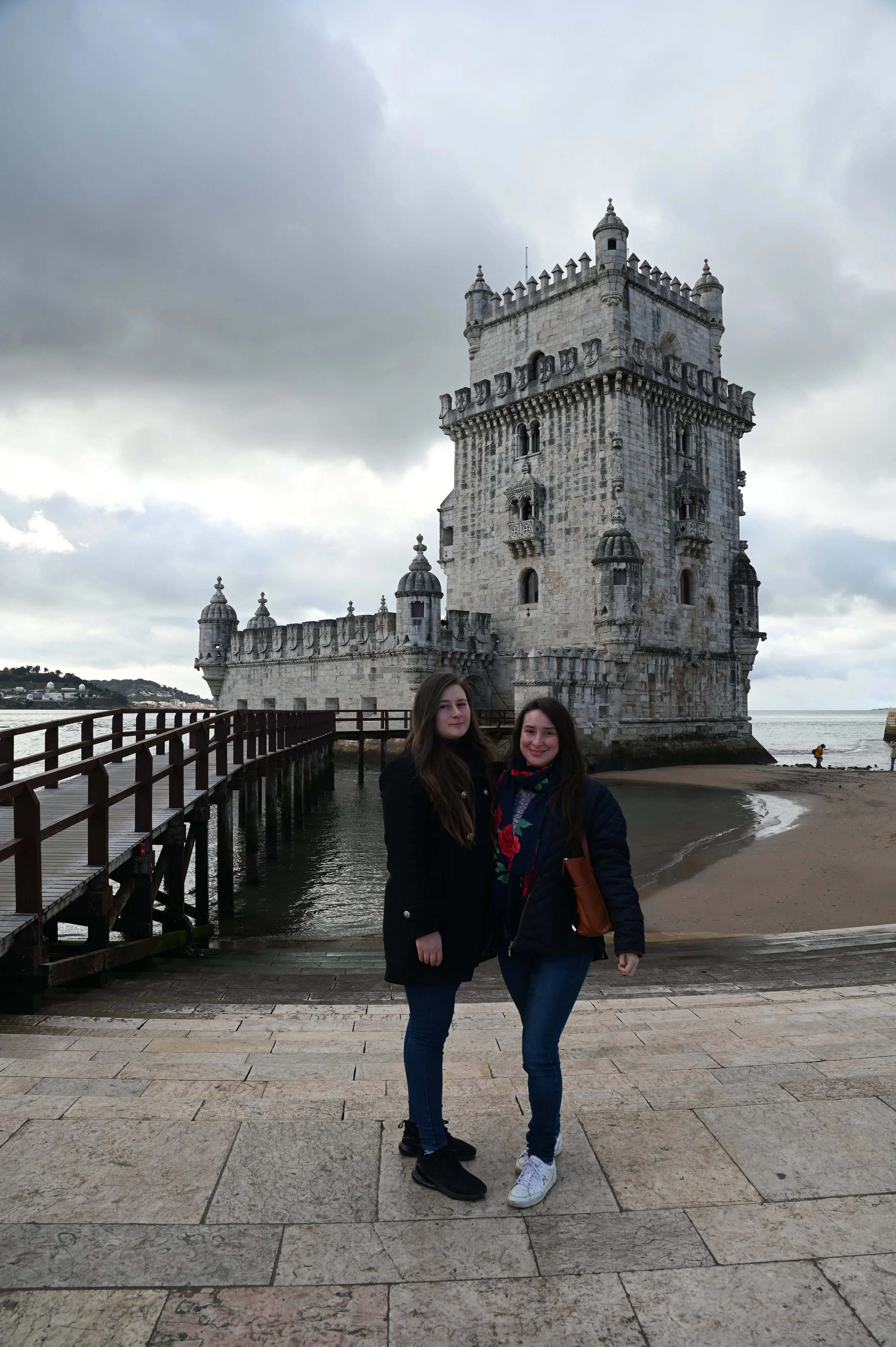 Two women standing in front of the Belem Tower, a historic fortress in Lisbon, Portugal, with cloudy sky in the background.