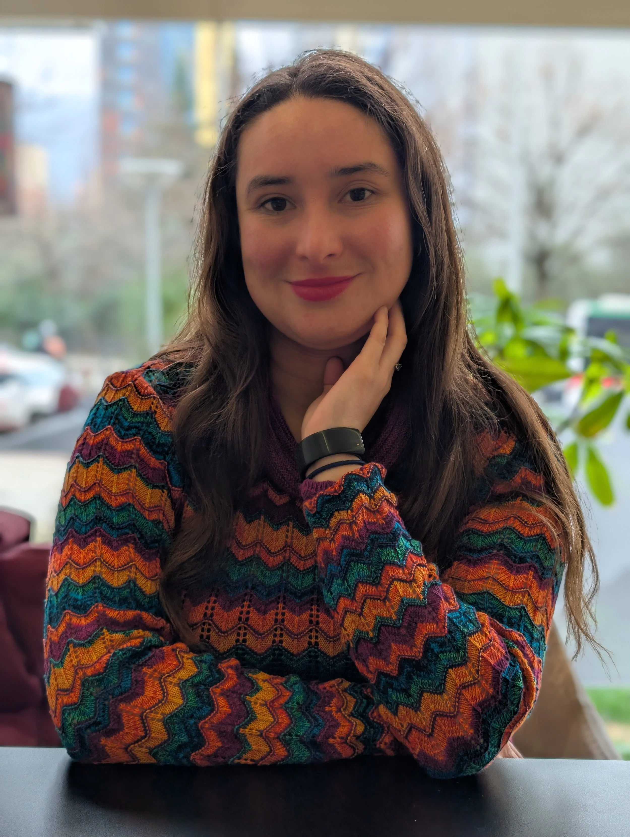 A woman with long brown hair, wearing a colorful patterned sweater, sitting at a table with her hand near her face, smiling softly, indoors with a window and outdoor trees in the background.