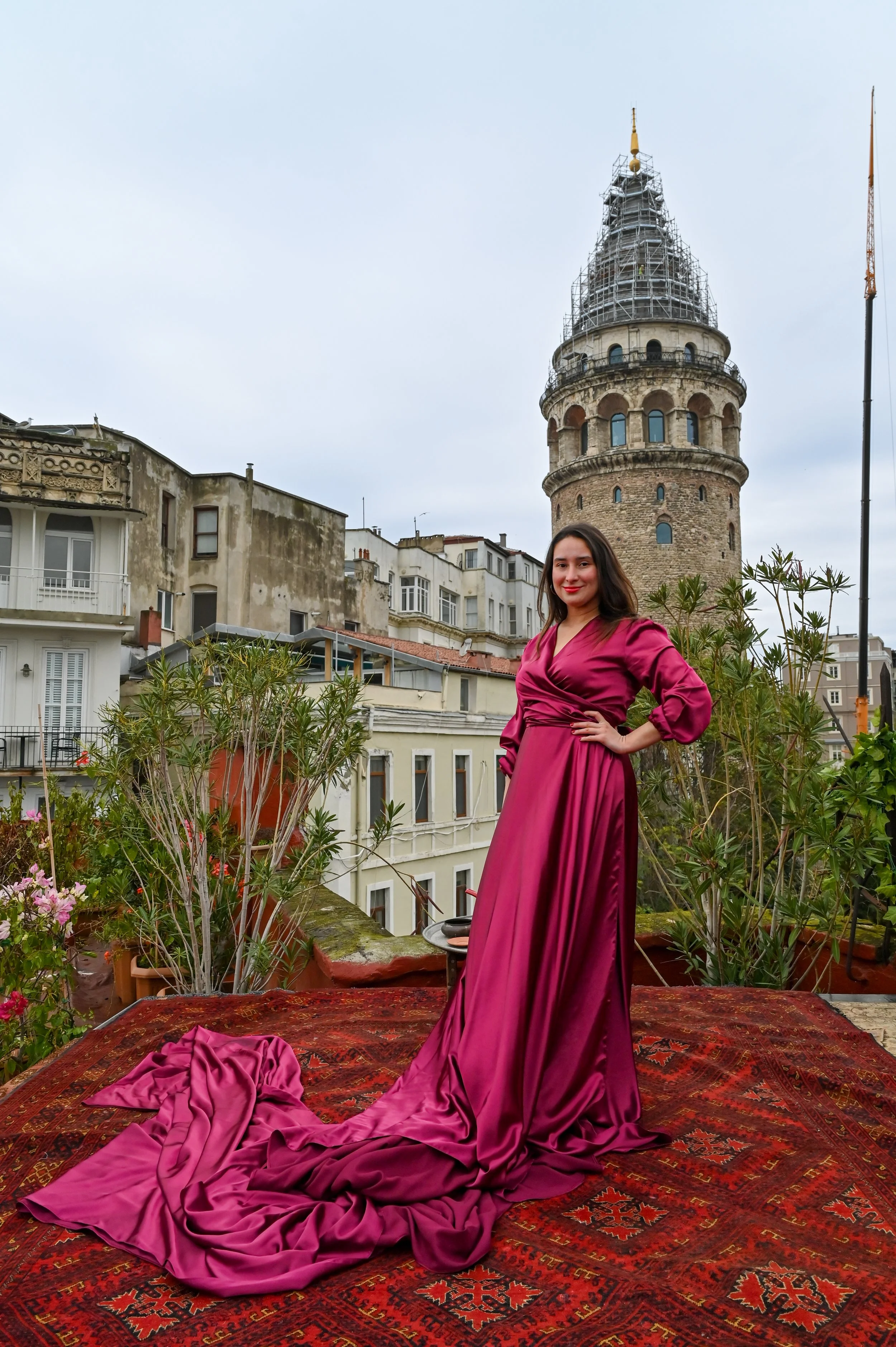 A woman in a long, magenta satin dress standing on a rooftop terrace with potted plants, surrounded by buildings and a historic tower in the background, cloudy sky.