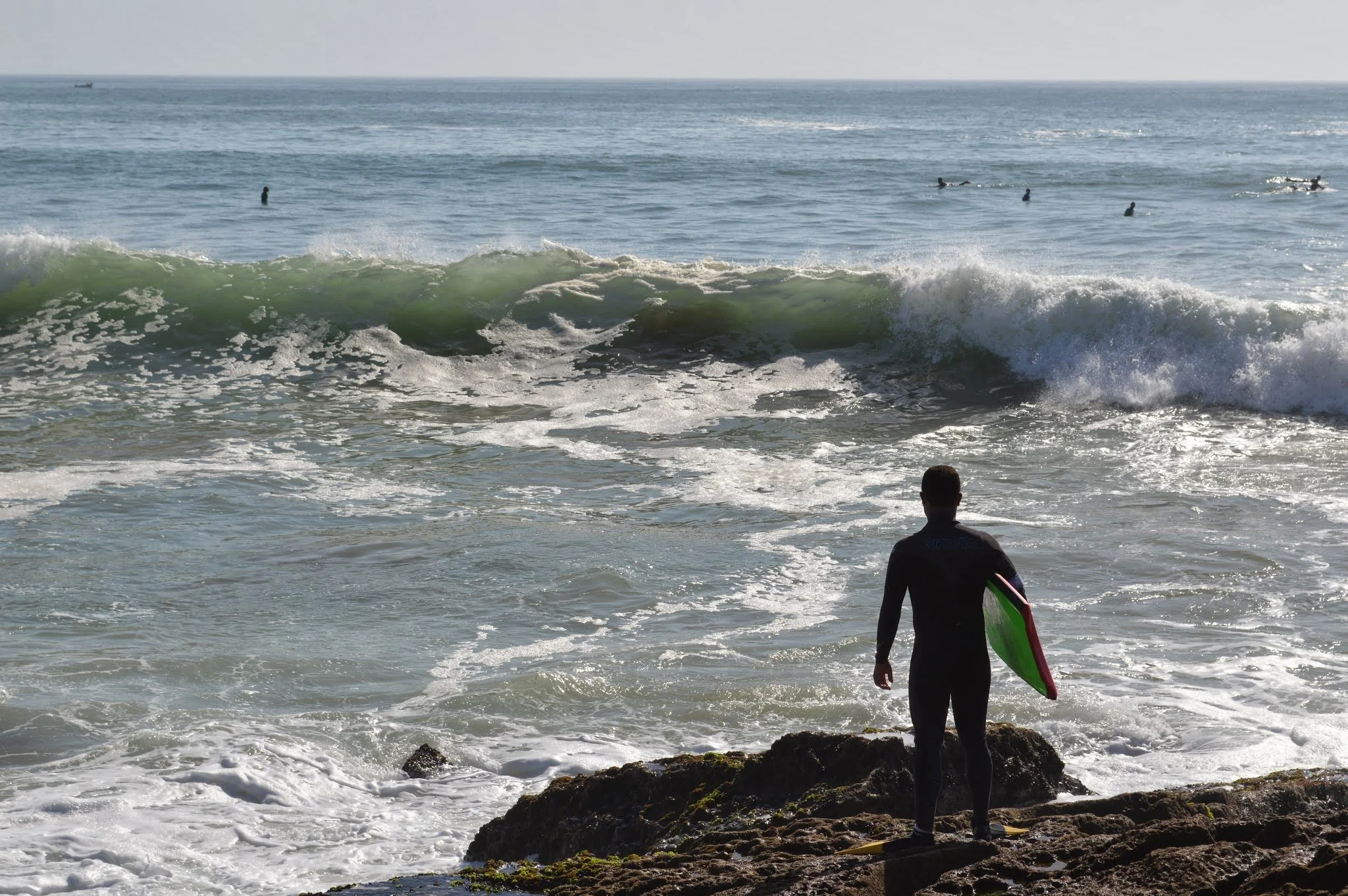A surfer in a wetsuit holding a green surfboard walking towards the ocean with waves and several people swimming in the water.