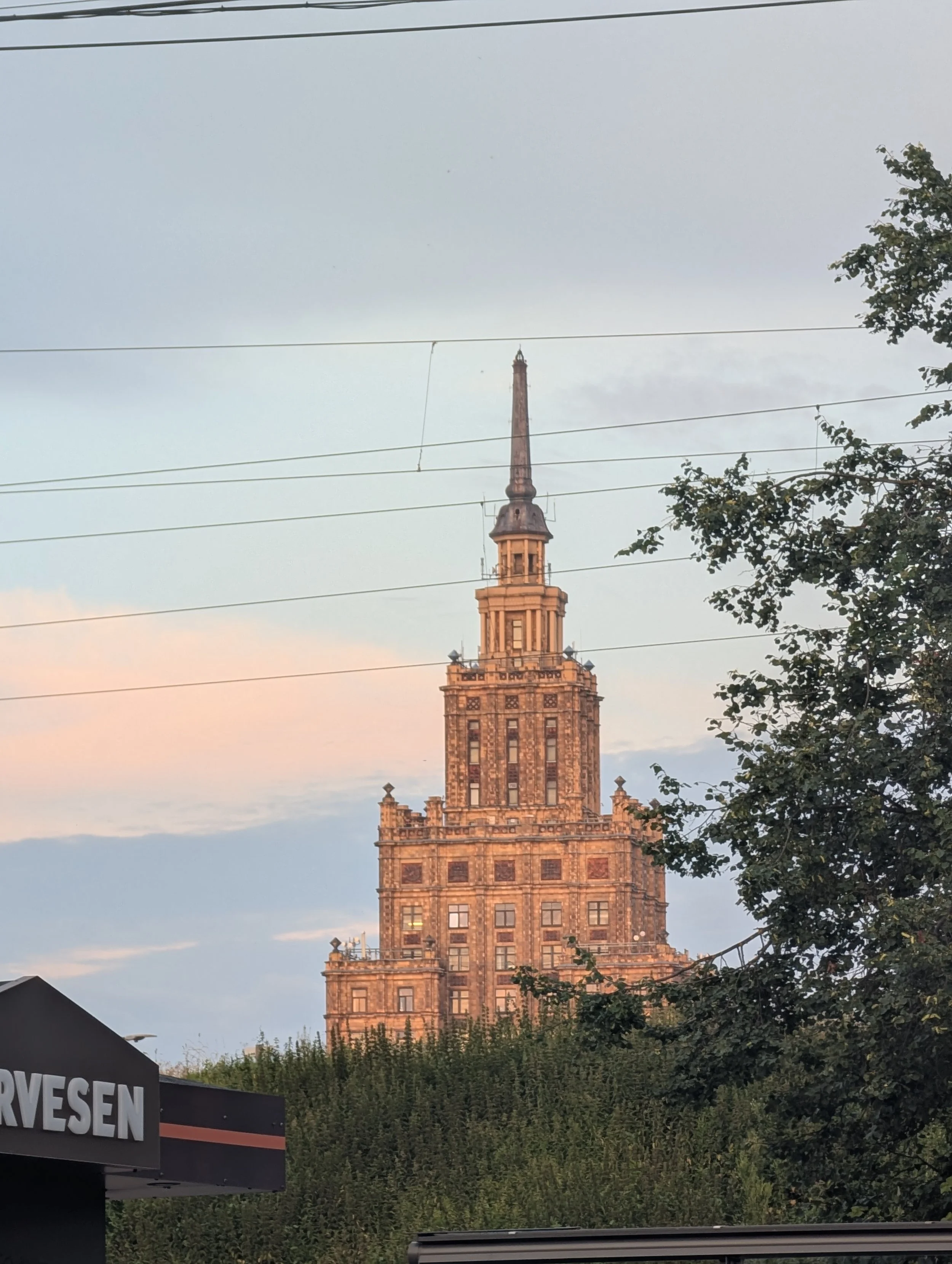 A tall historic building with a pointed spire, surrounded by trees, under a partly cloudy sky at sunset.