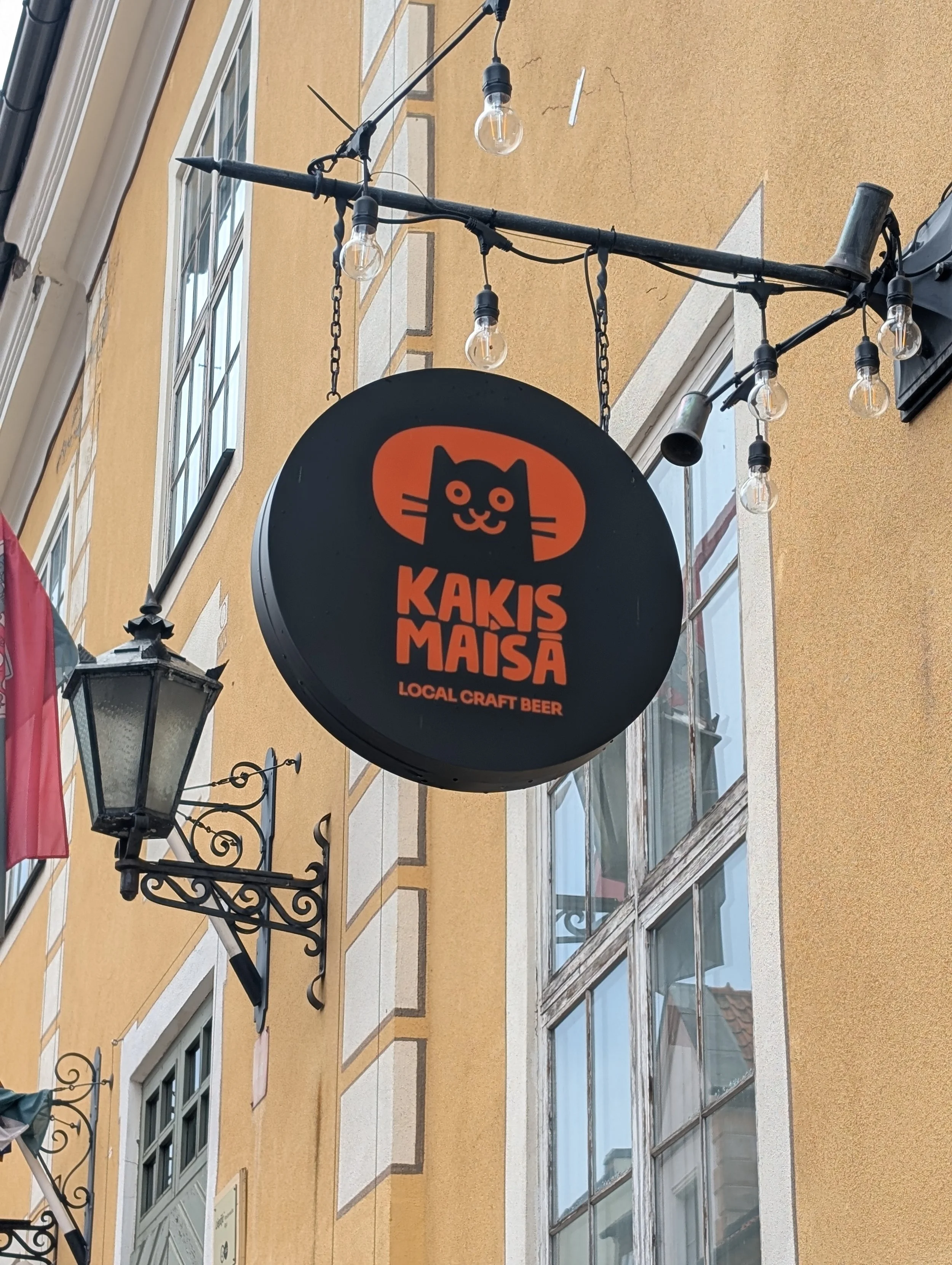 A circular sign with a black cat logo and the words 'Kakis Maisa' and 'Local Craft Beer' hanging on a black metal bracket outside a building with yellow walls and white-framed windows. String lights are hanging above the sign.