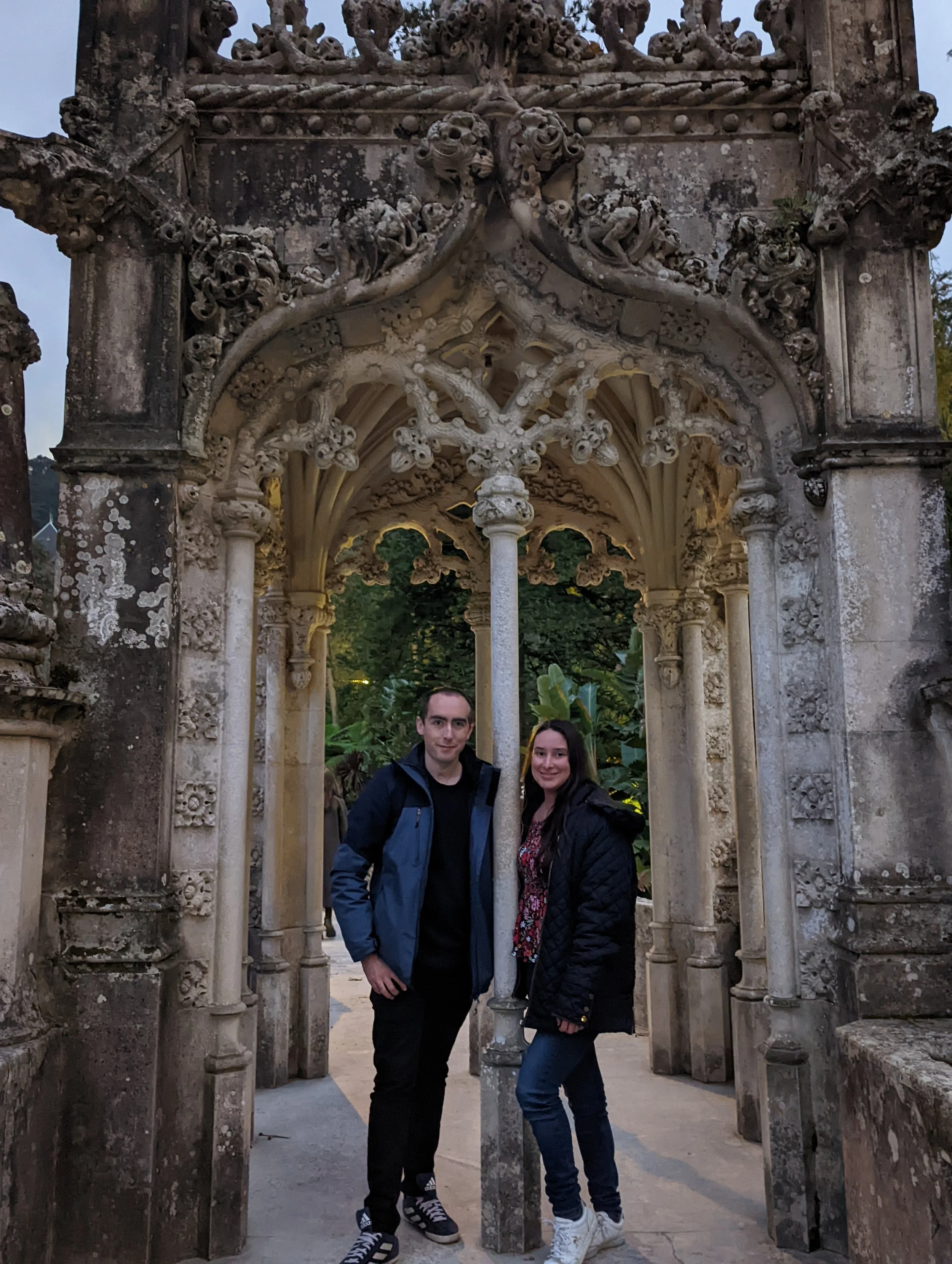 A man and woman standing together under a decorative stone archway with intricate carvings, outdoors in a garden area.