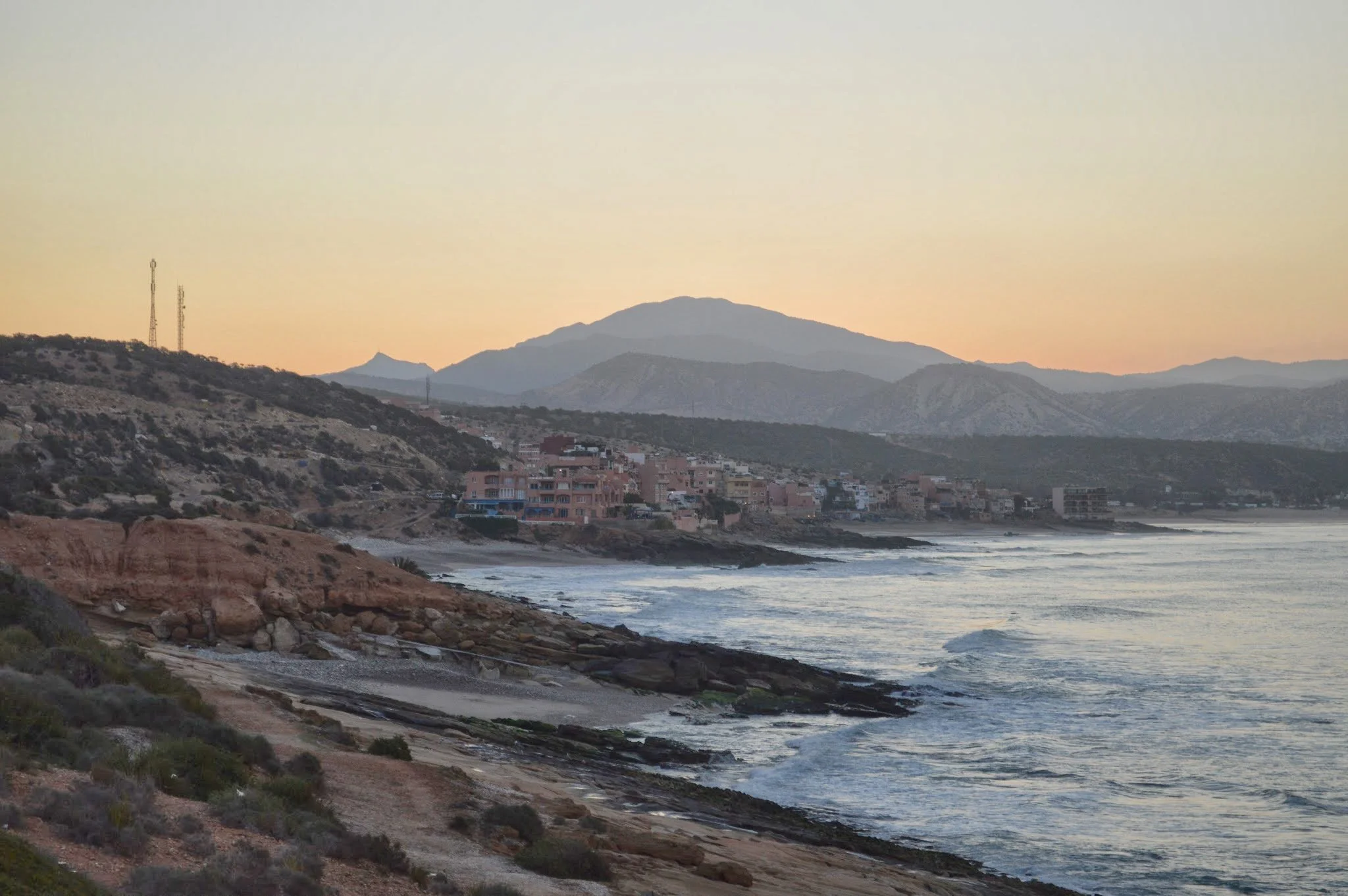 Scenic coastal view of mountains, beach, and buildings at sunset.