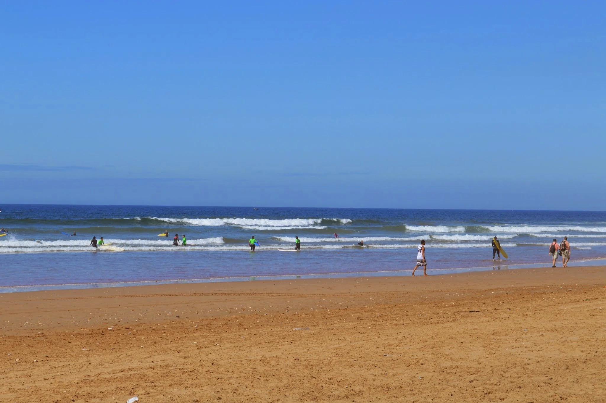 People enjoying the beach: some surfing, walking, and relaxing on the shore, under a blue sky with ocean waves.