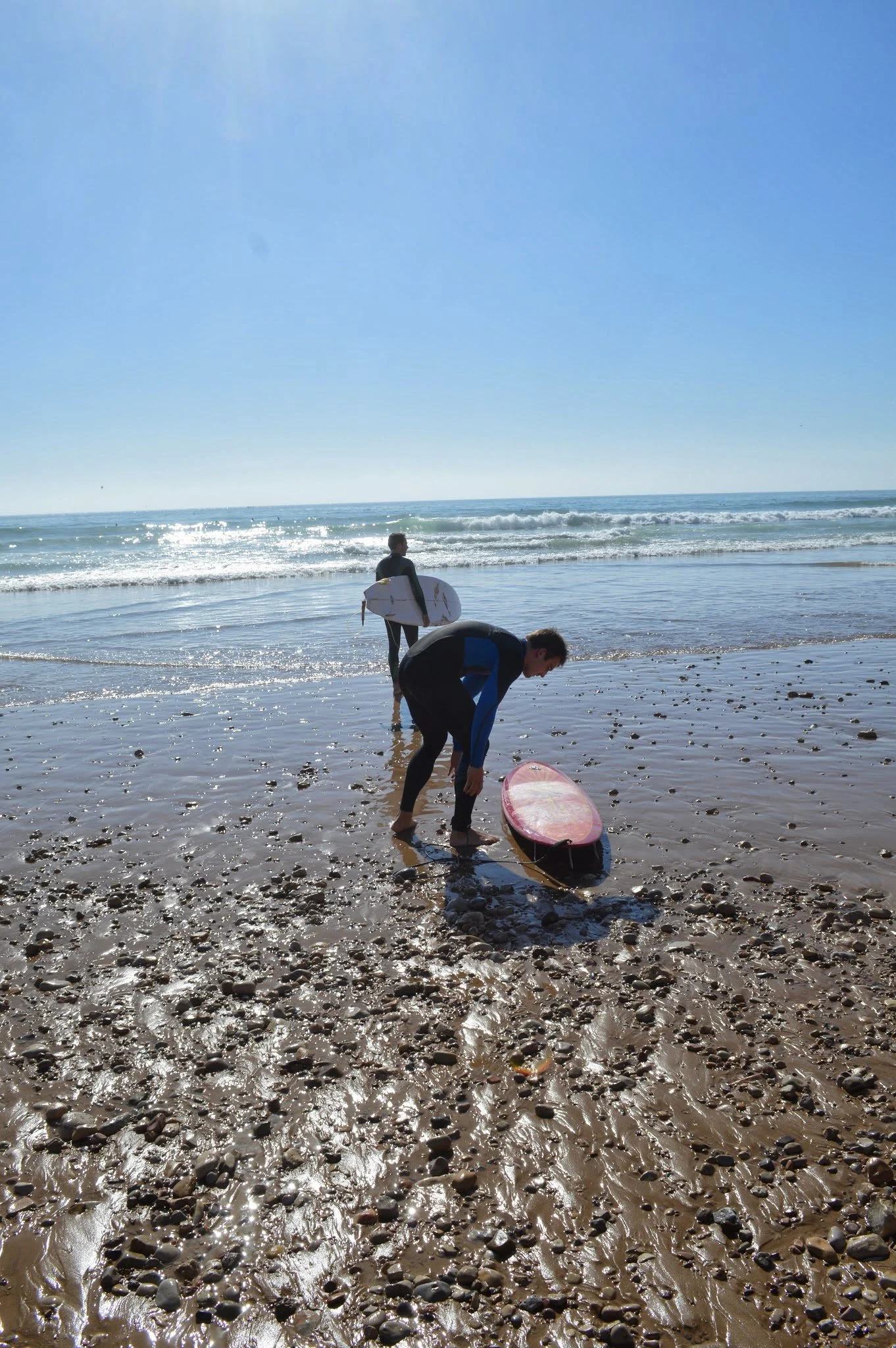 Two people preparing surfboards at the beach, one standing with a surfboard and the other bending over with a surfboard, with the ocean and sky in the background.