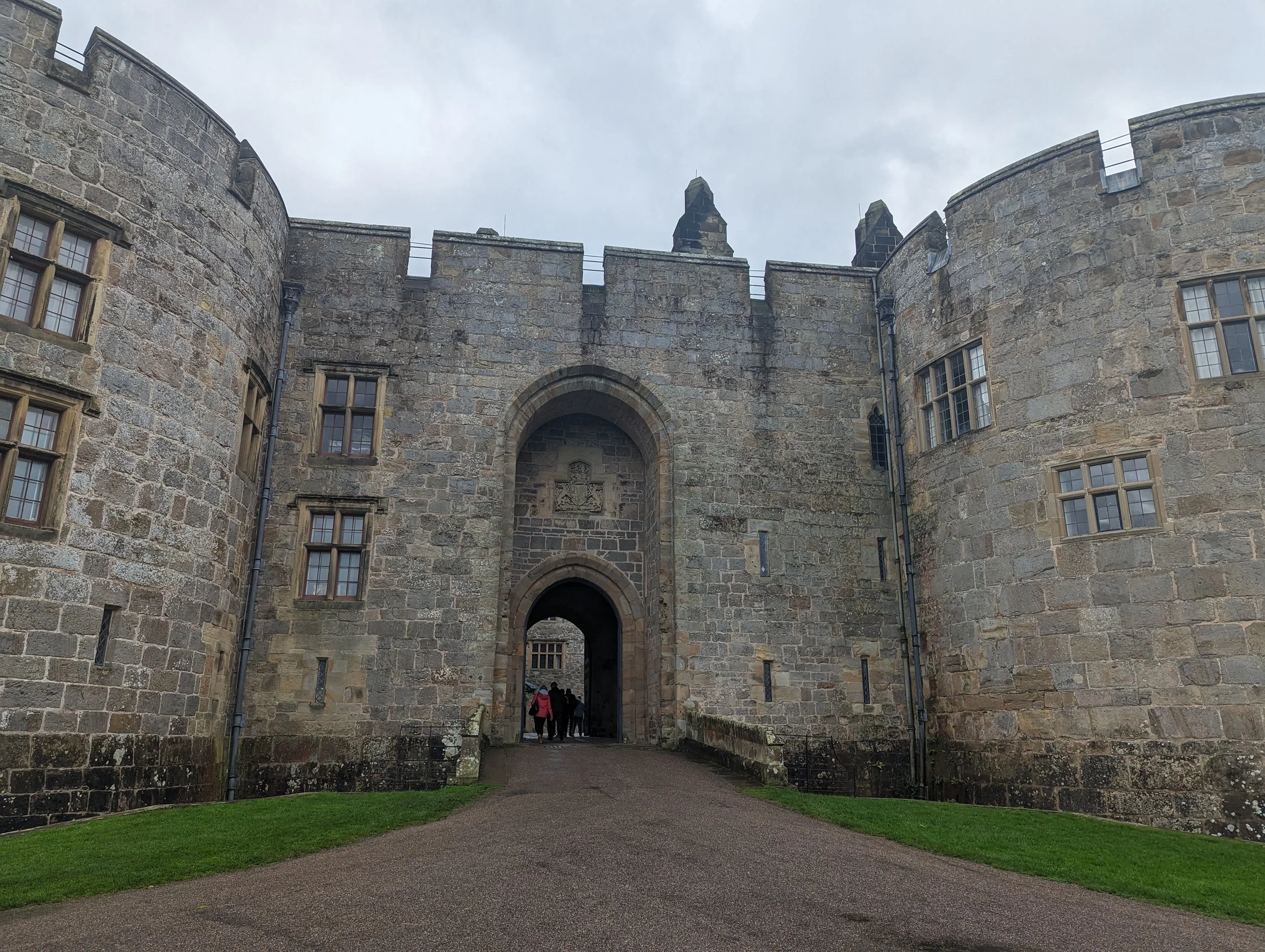 A stone castle or fortress with an arched entrance, multiple windows, and crenellated battlements on top, under a cloudy sky. Several people are walking through the entrance.