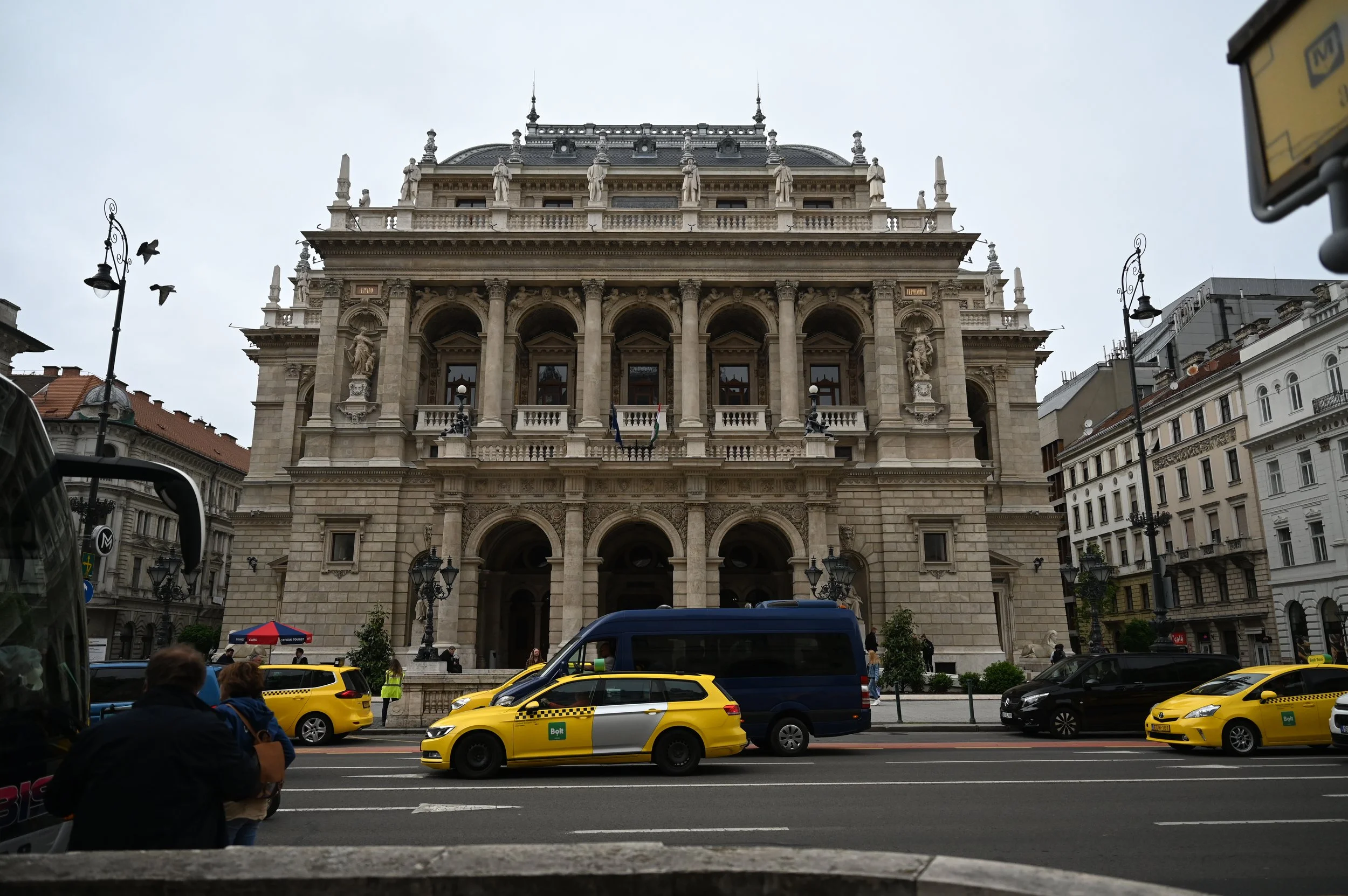 A grand, ornate building with classical architecture, including columns and sculptures, seen from the street. Yellow taxis and cars are in traffic, with pedestrians on the sidewalk.