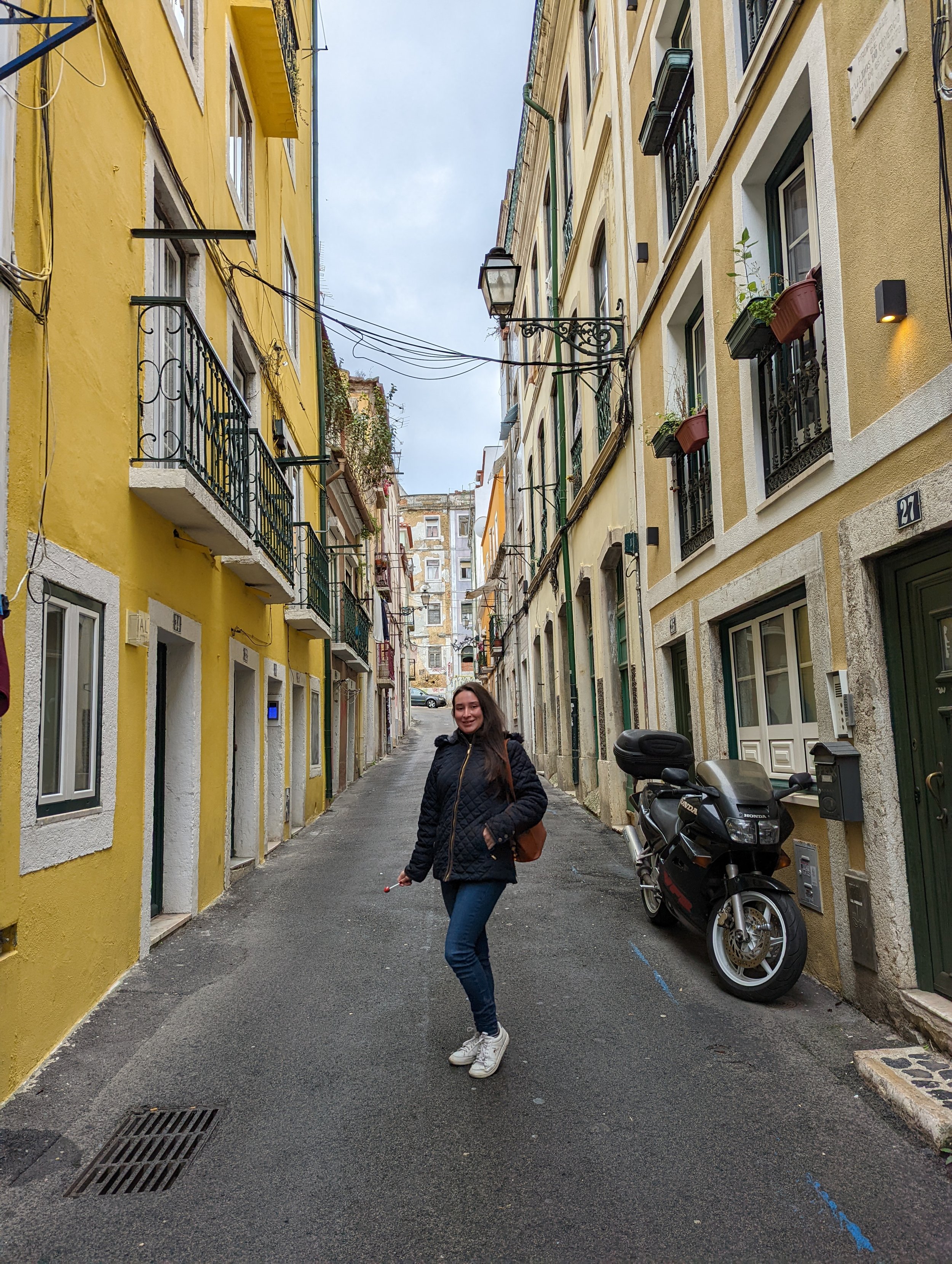 A woman in a black quilted jacket, jeans, and white sneakers standing in a narrow city street with yellow and beige buildings on either side. She is smiling and holding a small object in her right hand. A motorcycle is parked nearby, and the street a