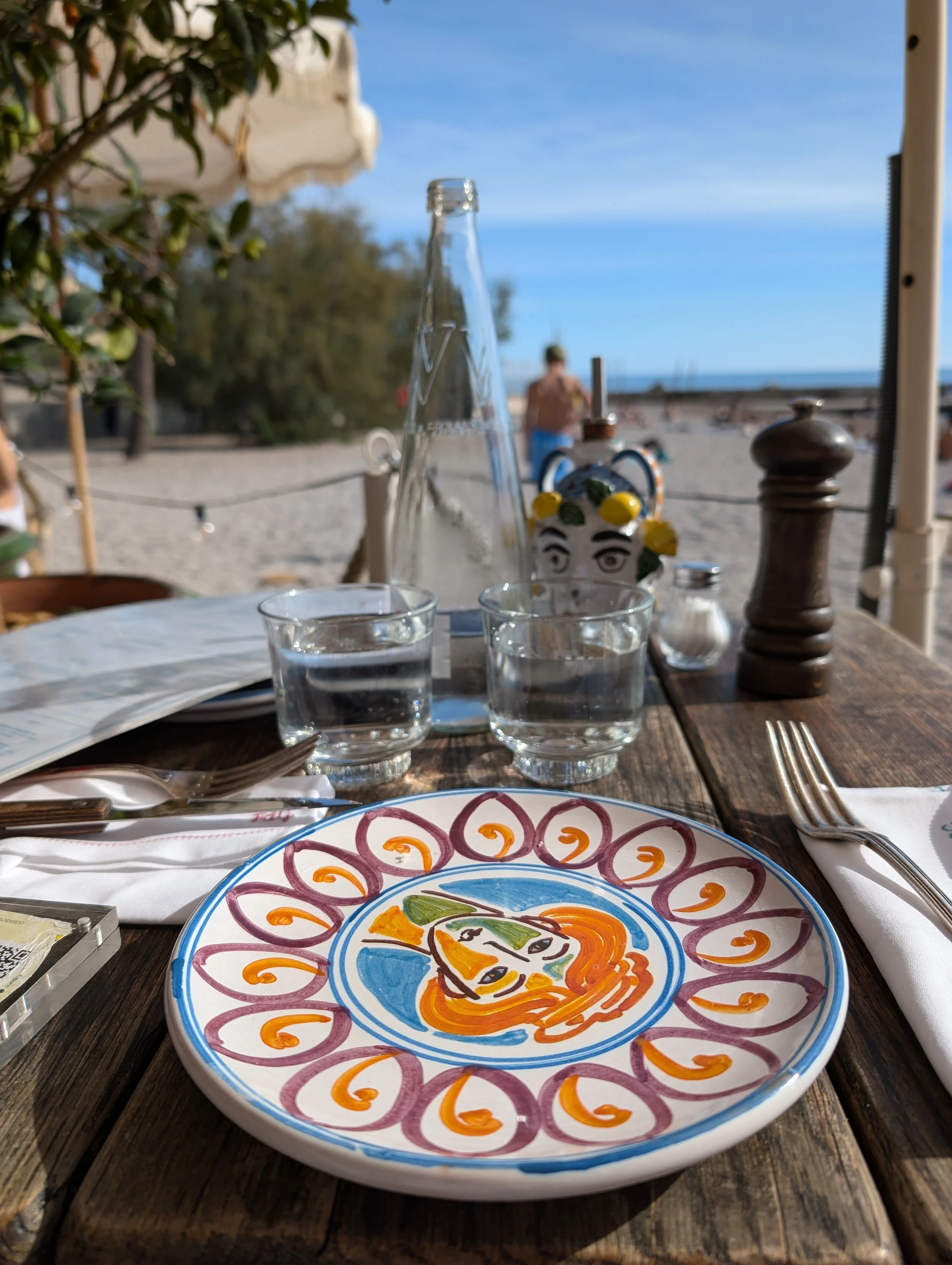 Colorful decorative plate with a stylized face design, set on a wooden table at an outdoor beachside setting.