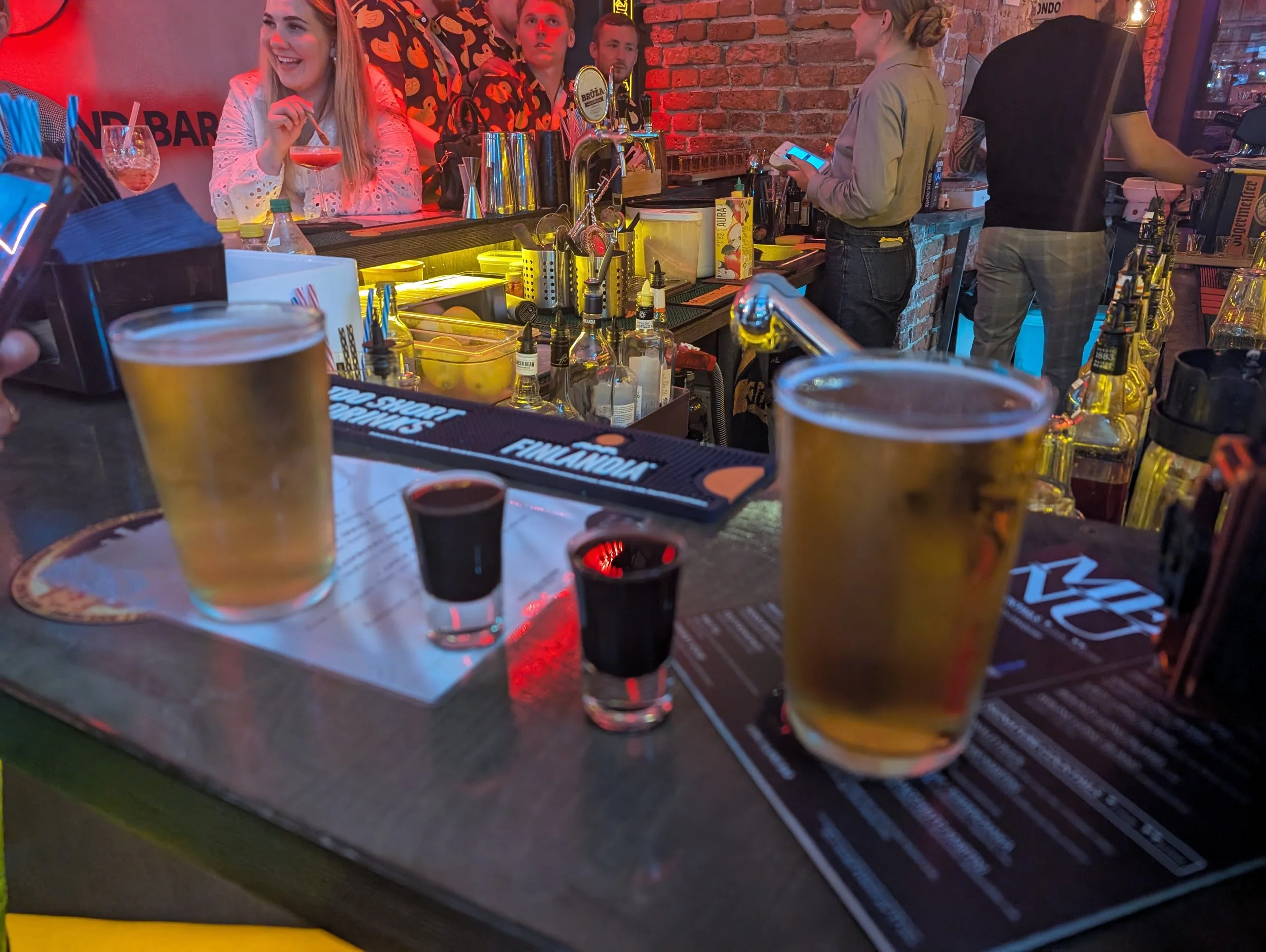 People at the bar in a lively social setting with drinks on the counter, exposed brick walls, and colorful lighting.