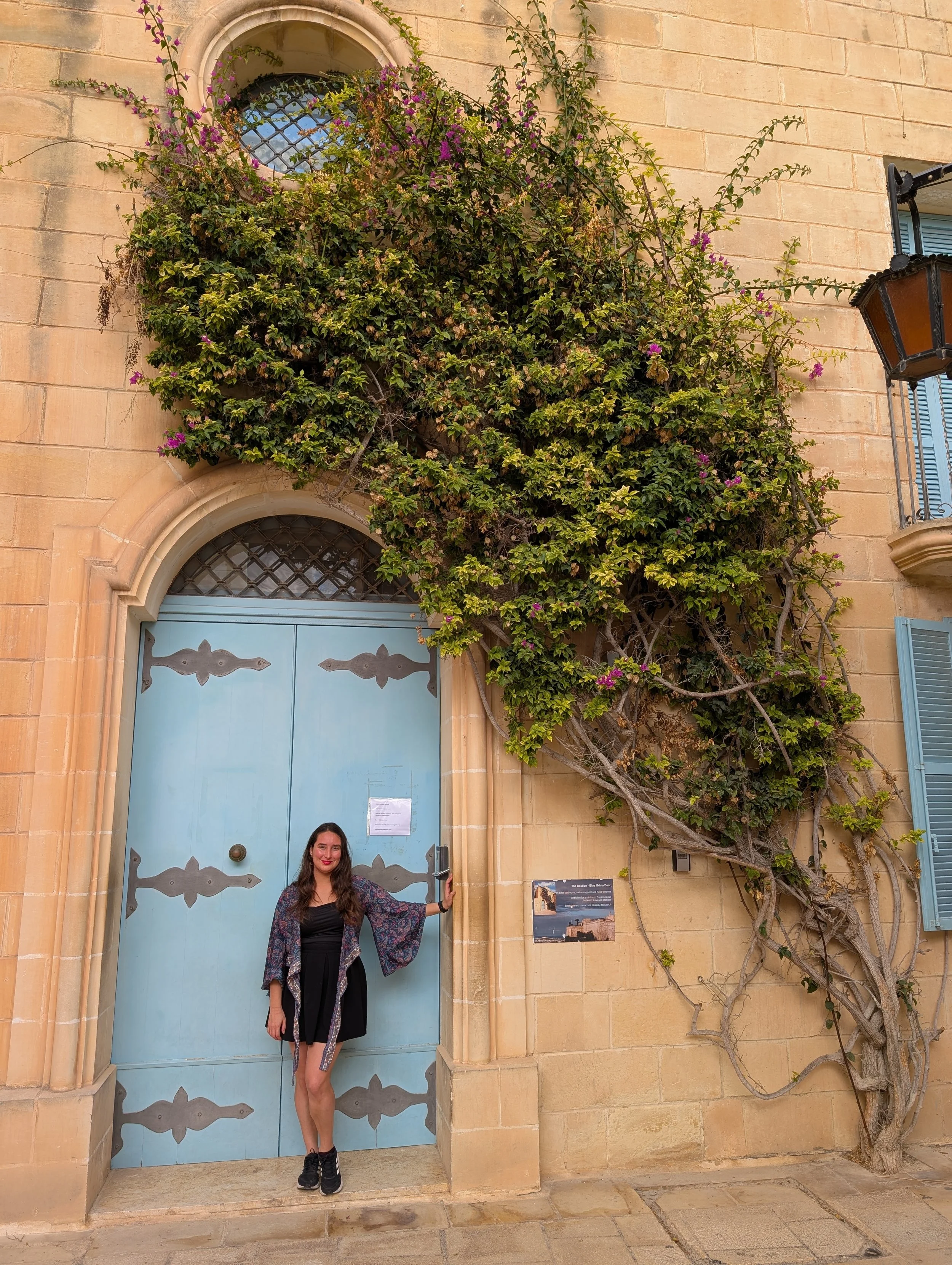 A woman with long dark hair, wearing black shoes, a black dress, and a floral shawl, standing in front of a large blue door with decorative metalwork, holding the door handle. The building has a rustic stone exterior with climbing green and purple fl