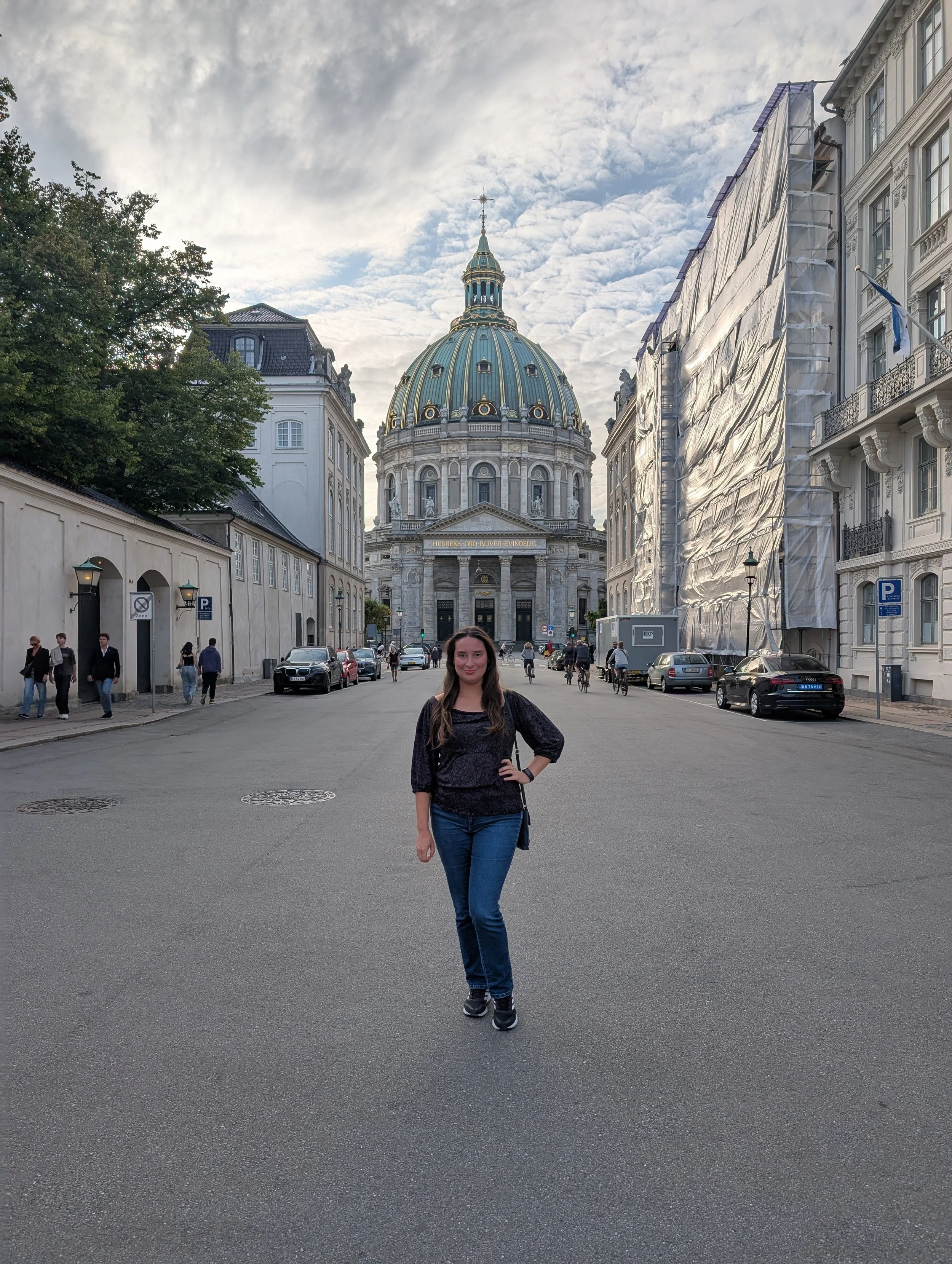 A woman standing in the middle of a street with historical buildings and a domed church in the background, under a cloudy sky.