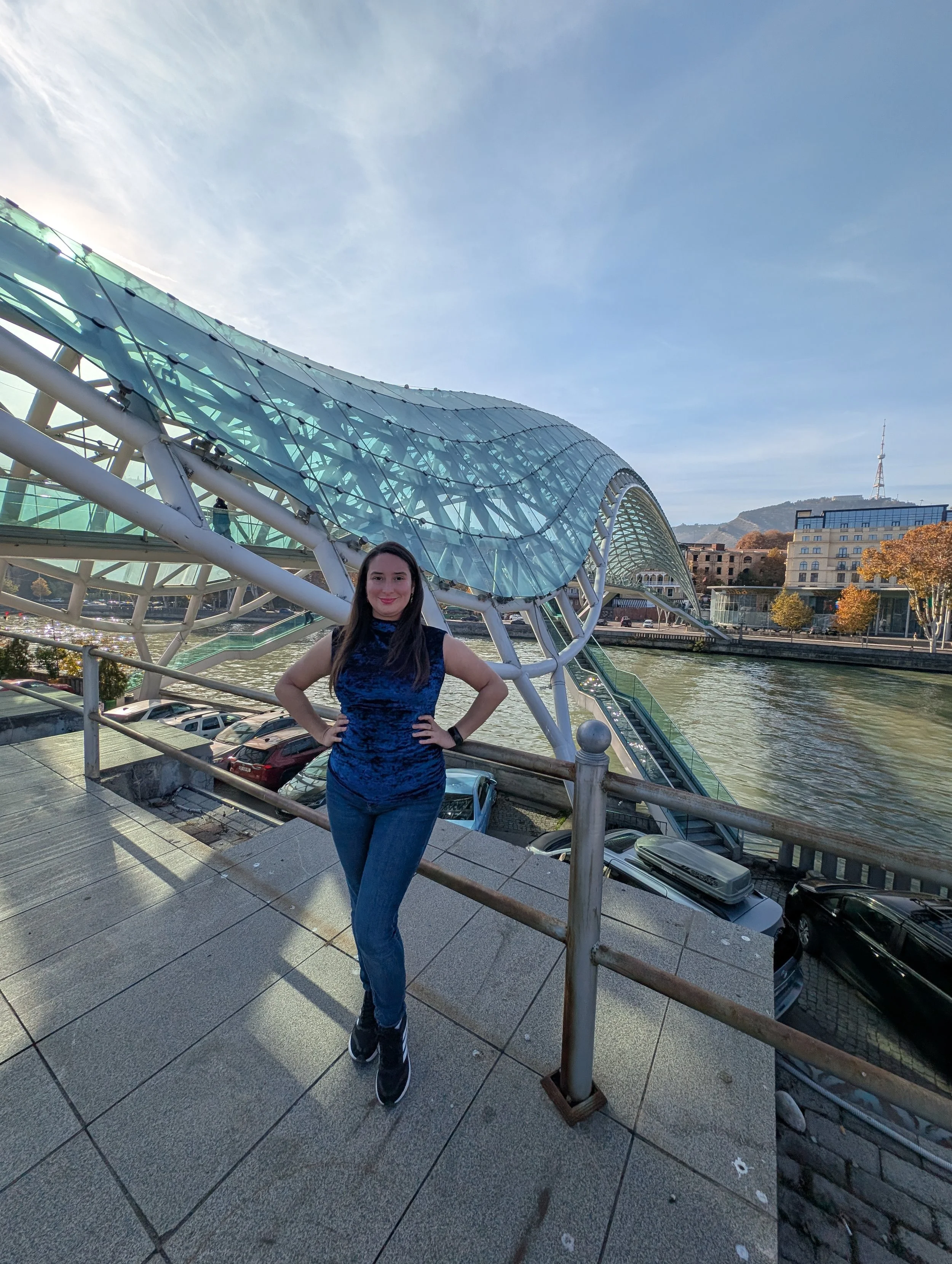 A woman standing on a bridge with metal railings over a river, with blue sky, modern architectural structure, and city buildings in the background during daytime.