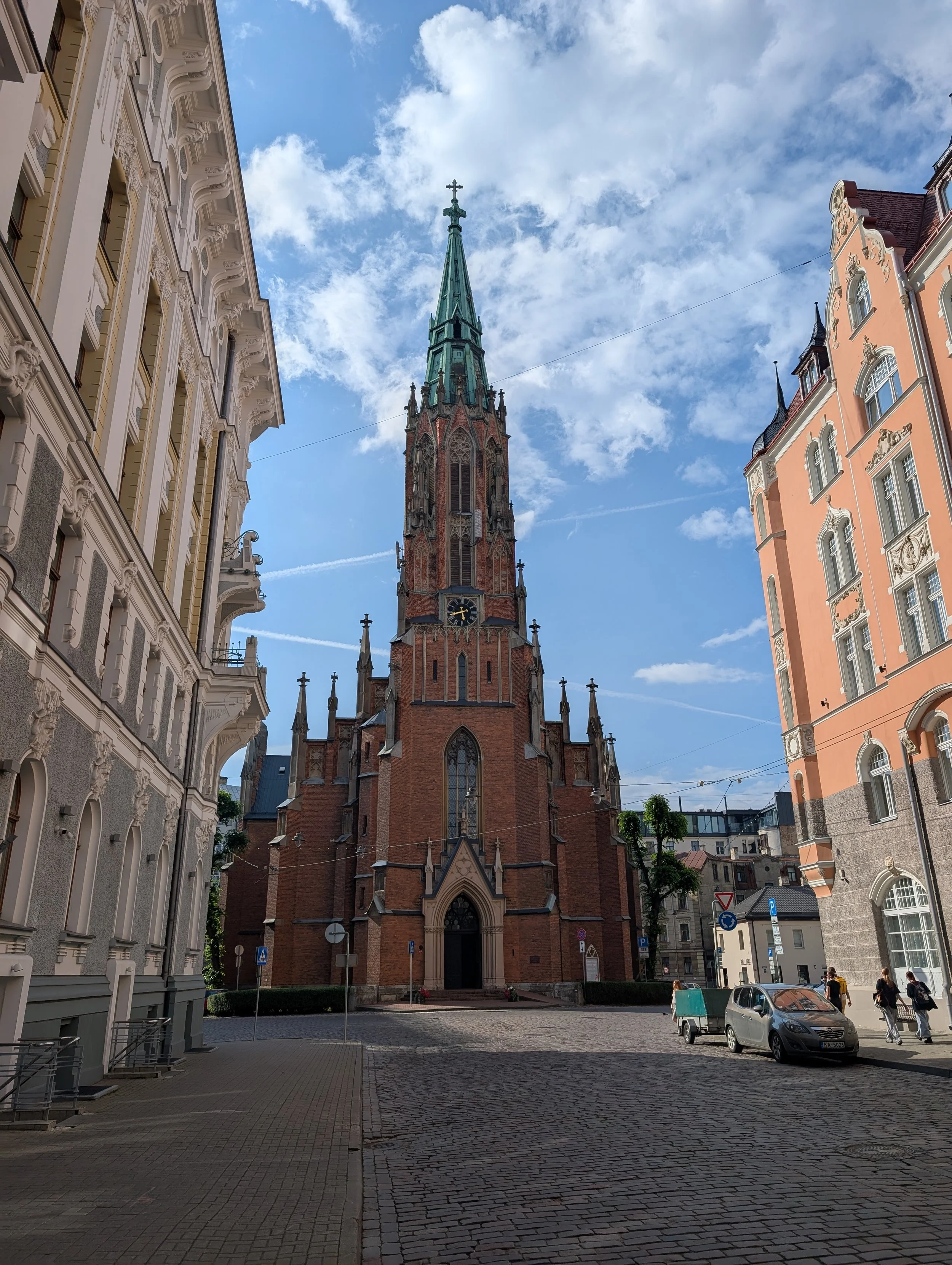 View of a historic church with a tall steeple, surrounded by ornate buildings on a cobblestone street, under a partly cloudy sky.