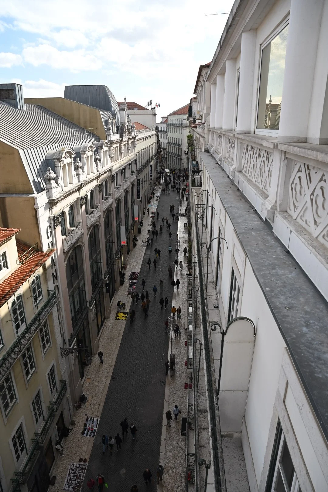 A busy pedestrian street view from an elevated position, showing people walking and street vendors selling items, lined with classic European-style buildings with large windows and ornate facades.
