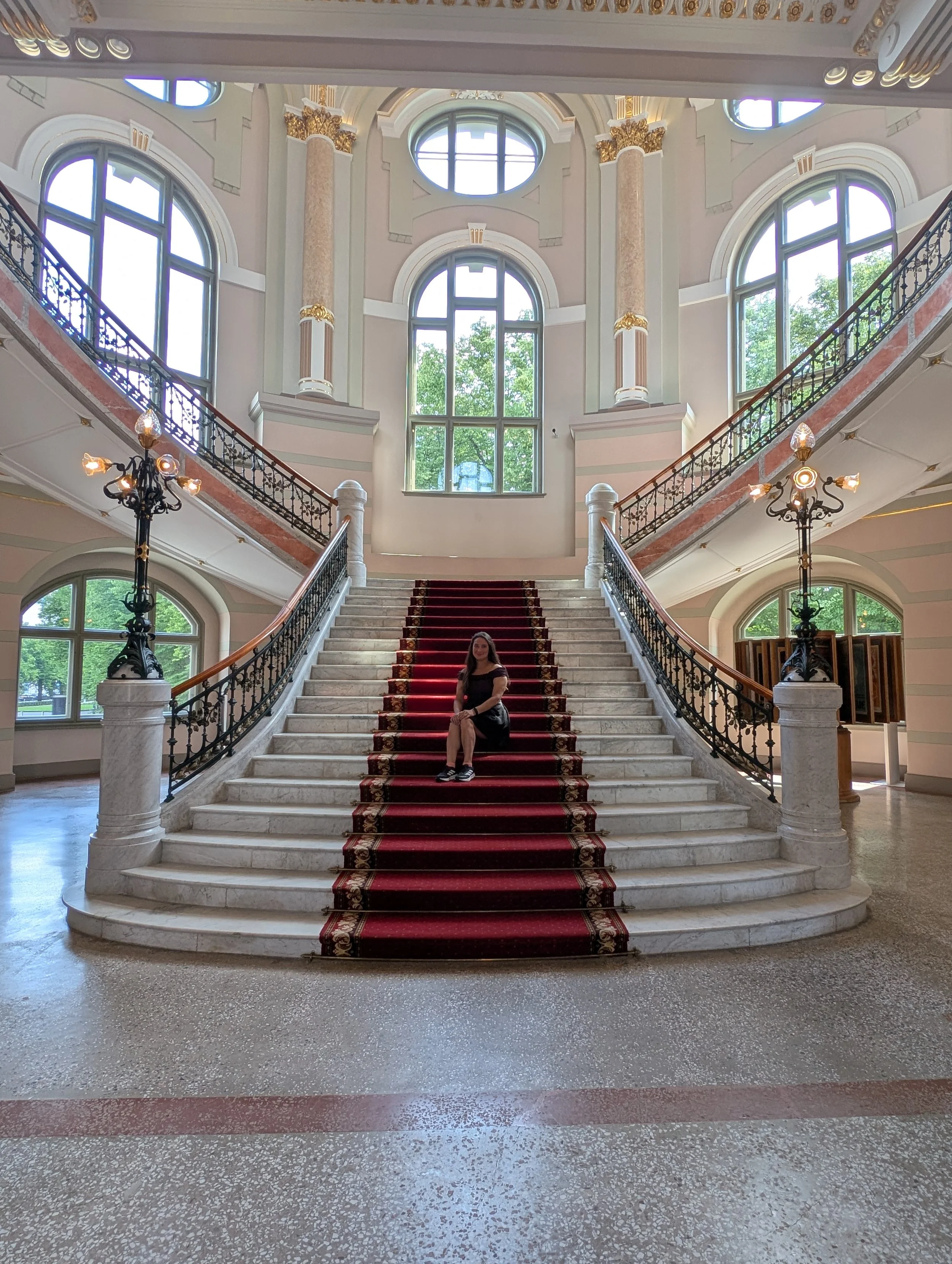 Interior view of an elegant palace staircase with a red carpet, ornate black iron railings, marble pillars with gold accents, large arched windows, and chandeliers, with a woman sitting on the stairs.