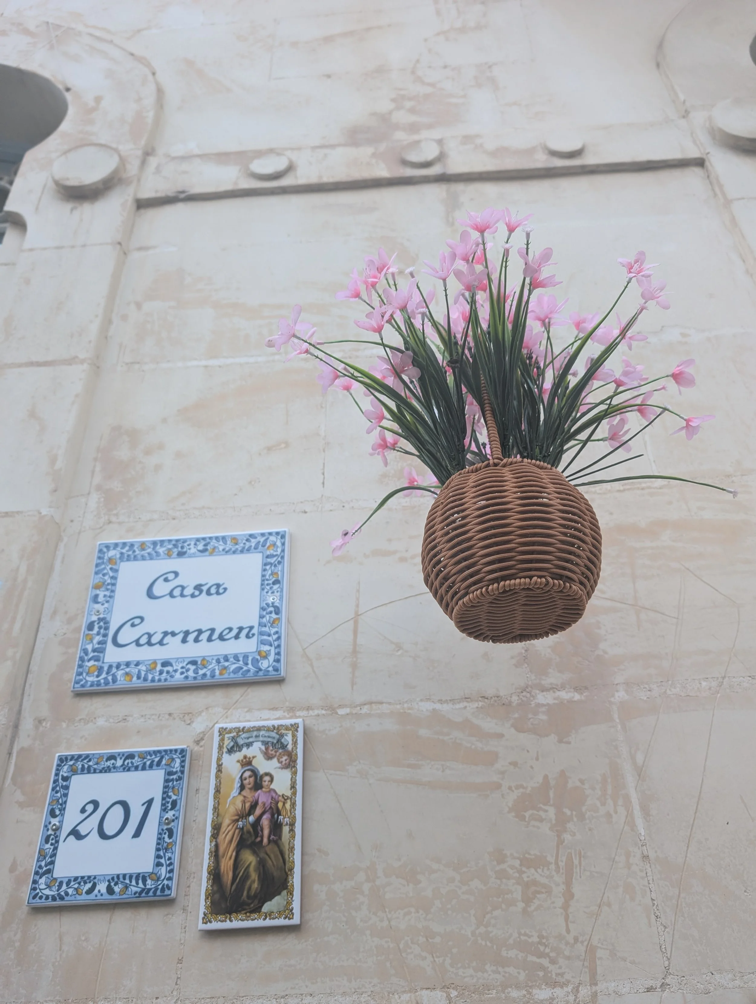 A wicker basket mounted on the wall containing pink flowers. Below are two decorative ceramic tiles with blue and white patterns, with one featuring the number 201 and the other showing religious imagery.