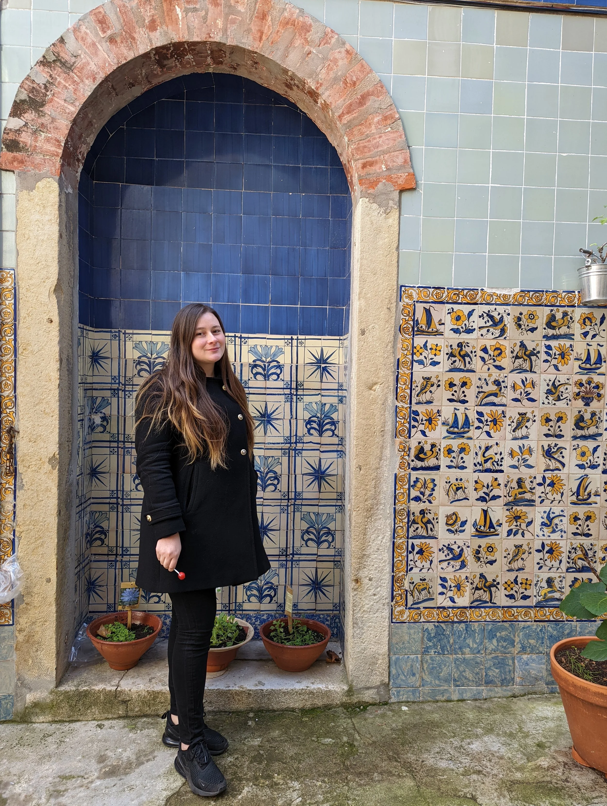 A woman with long brown hair wearing a black coat and black sneakers standing next to an outdoor tiled wall with decorative patterns and blue tiles, surrounded by potted plants, on a concrete surface.