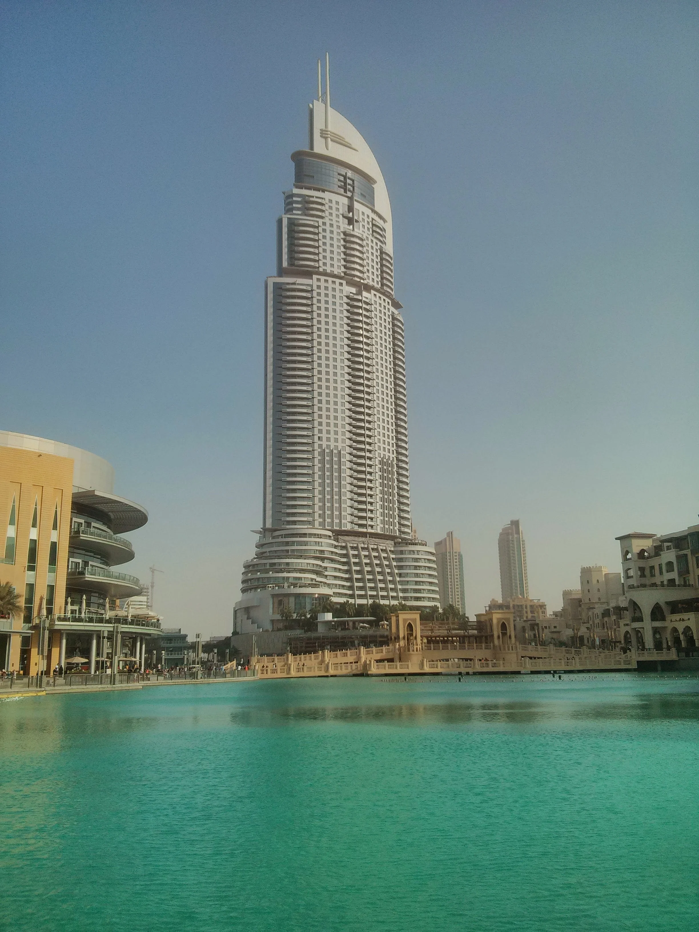 A modern high-rise building with a distinctive curved design and multiple balconies, situated near a body of water with a marina and additional buildings in a cityscape.