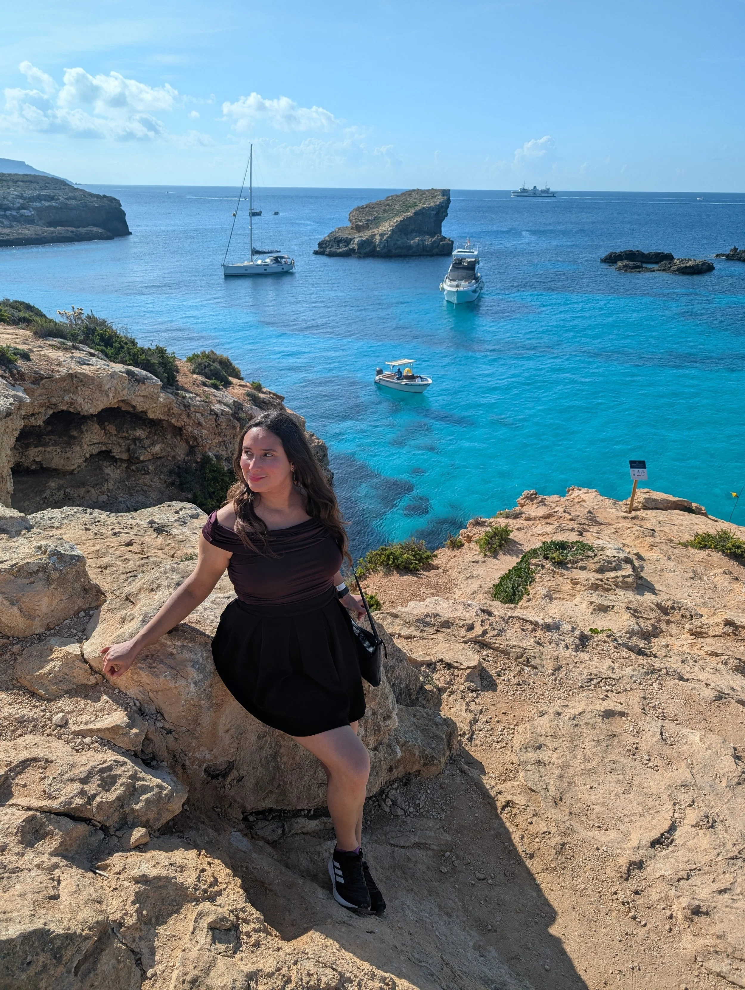 A woman in a black skirt and black shoes posing on rocky terrain near the ocean with boats and a large rock formation in the background, under a partly cloudy sky.
