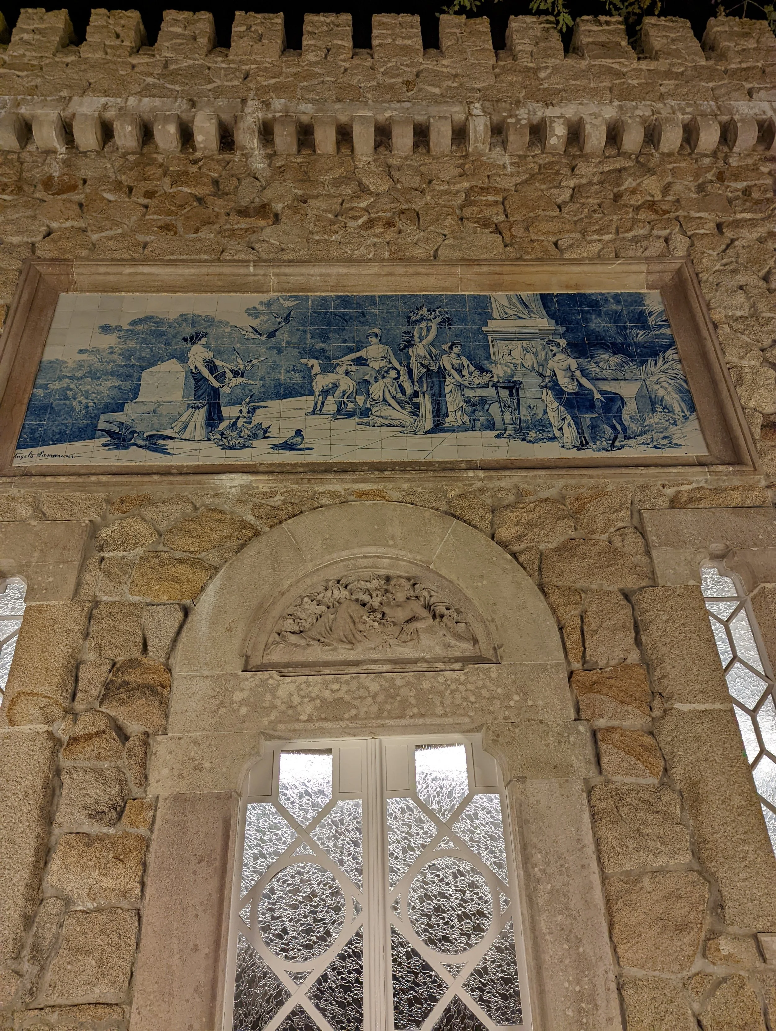 A mural painted on tiles depicts a historical scene with people and animals, framed by a stone wall and a decorative white window with lattice and textured glass, located above an arched stone window with intricate carvings.