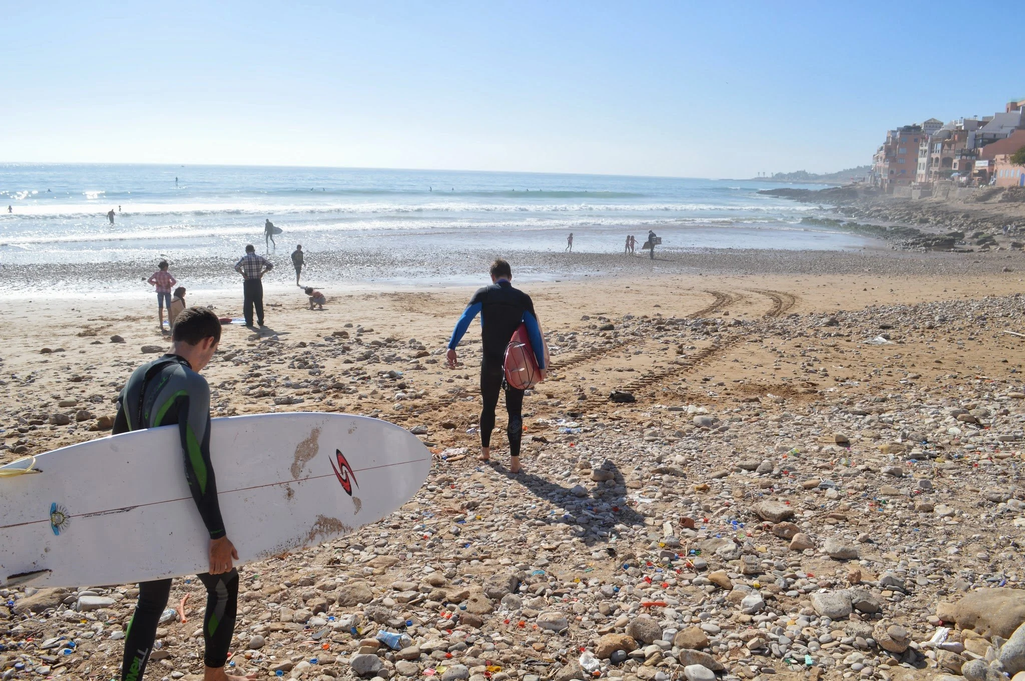 People at a beach with a rocky and sandy shoreline, some holding surfing boards, watching the waves under a clear blue sky with buildings on the right side.