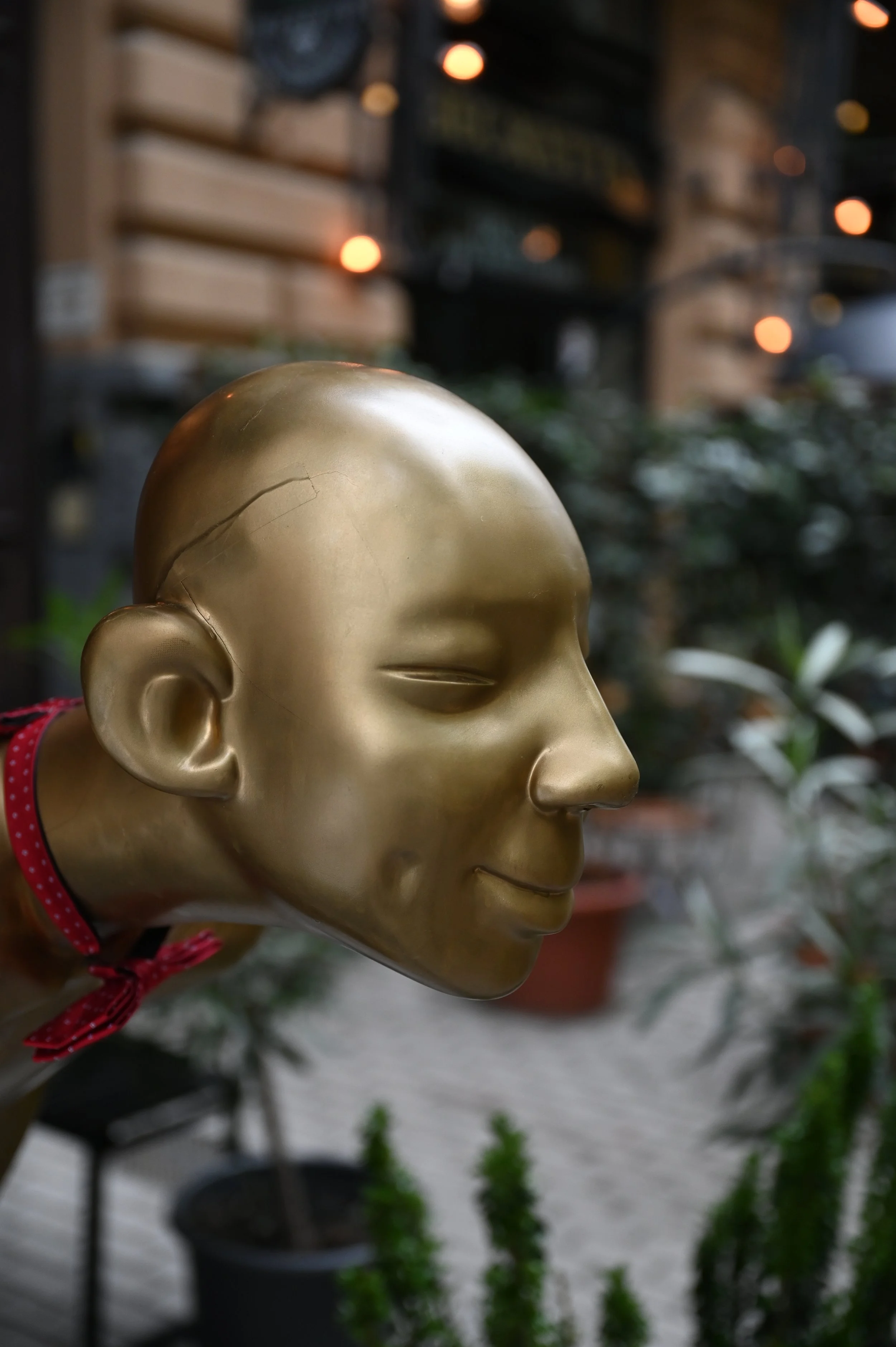 A golden Buddha face sculpture with closed eyes, set against a blurred background with orange lights and greenery.