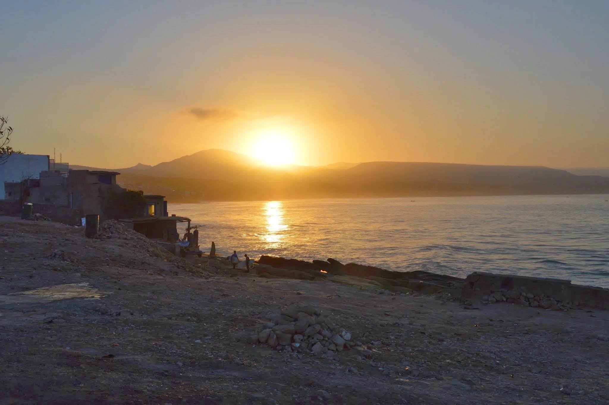 Sunset over a body of water with hills in the distance and a small town along the shoreline. There are buildings on the left side and a few people sitting on rocks in the foreground.