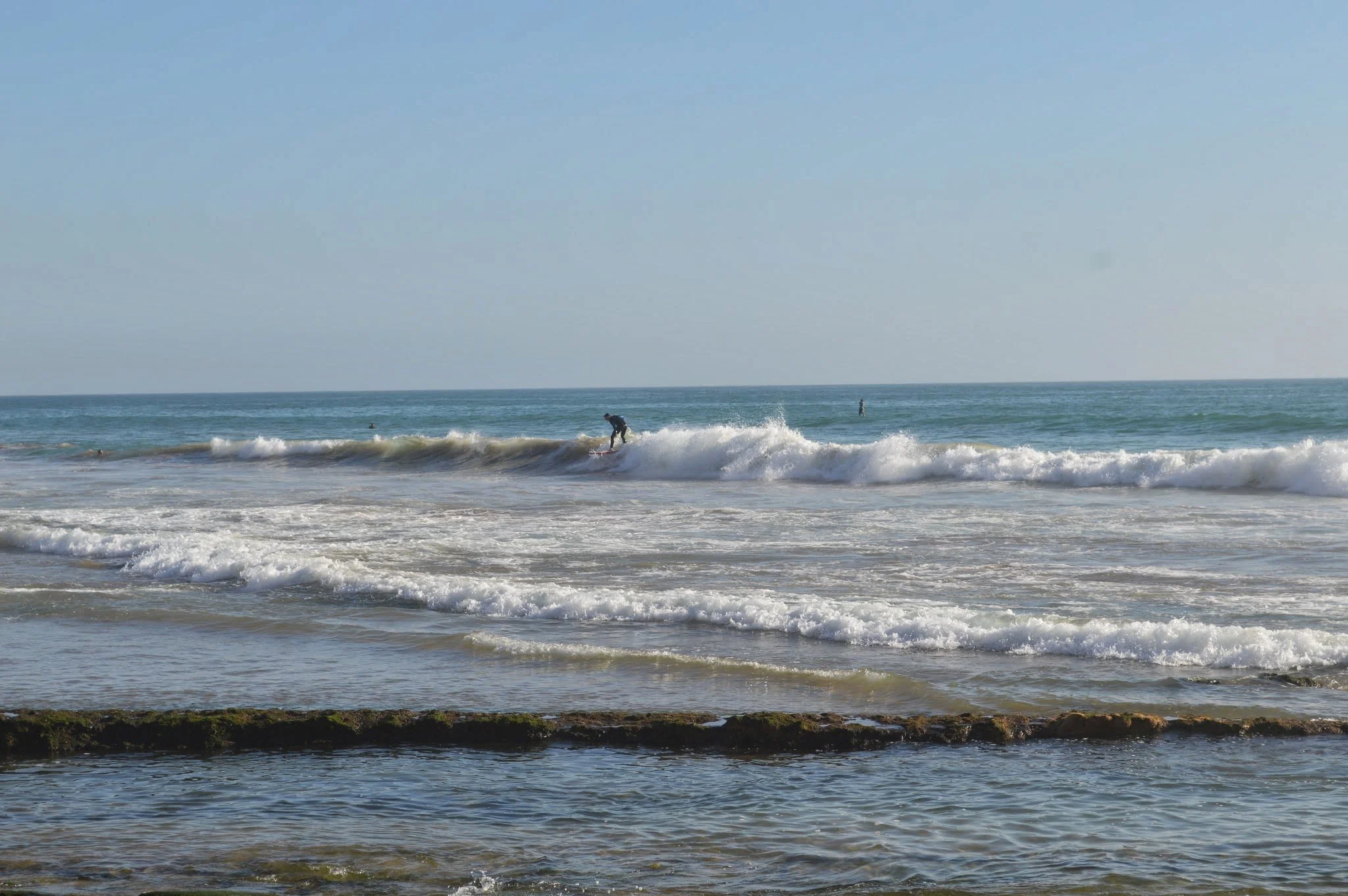 A beach scene with waves crashing onto the shore, people surfing in the water, and a clear blue sky overhead.