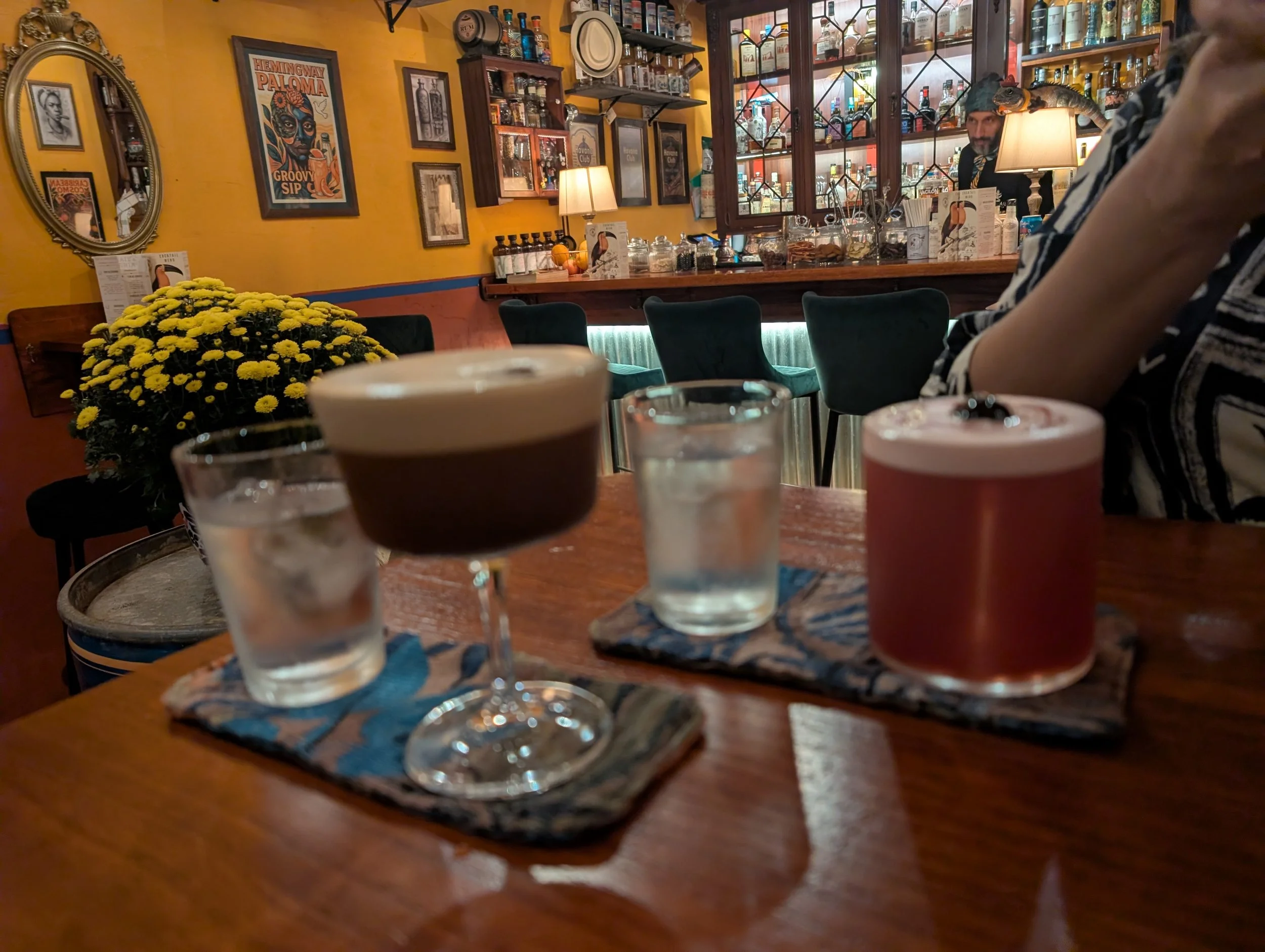 A cozy bar with yellow walls decorated with framed artwork and posters. A large mirror reflects part of the room. The bar counter is visible with liquor bottles, jars of garnishes, and bar tools. A bartender is behind the bar. In the foreground, ther