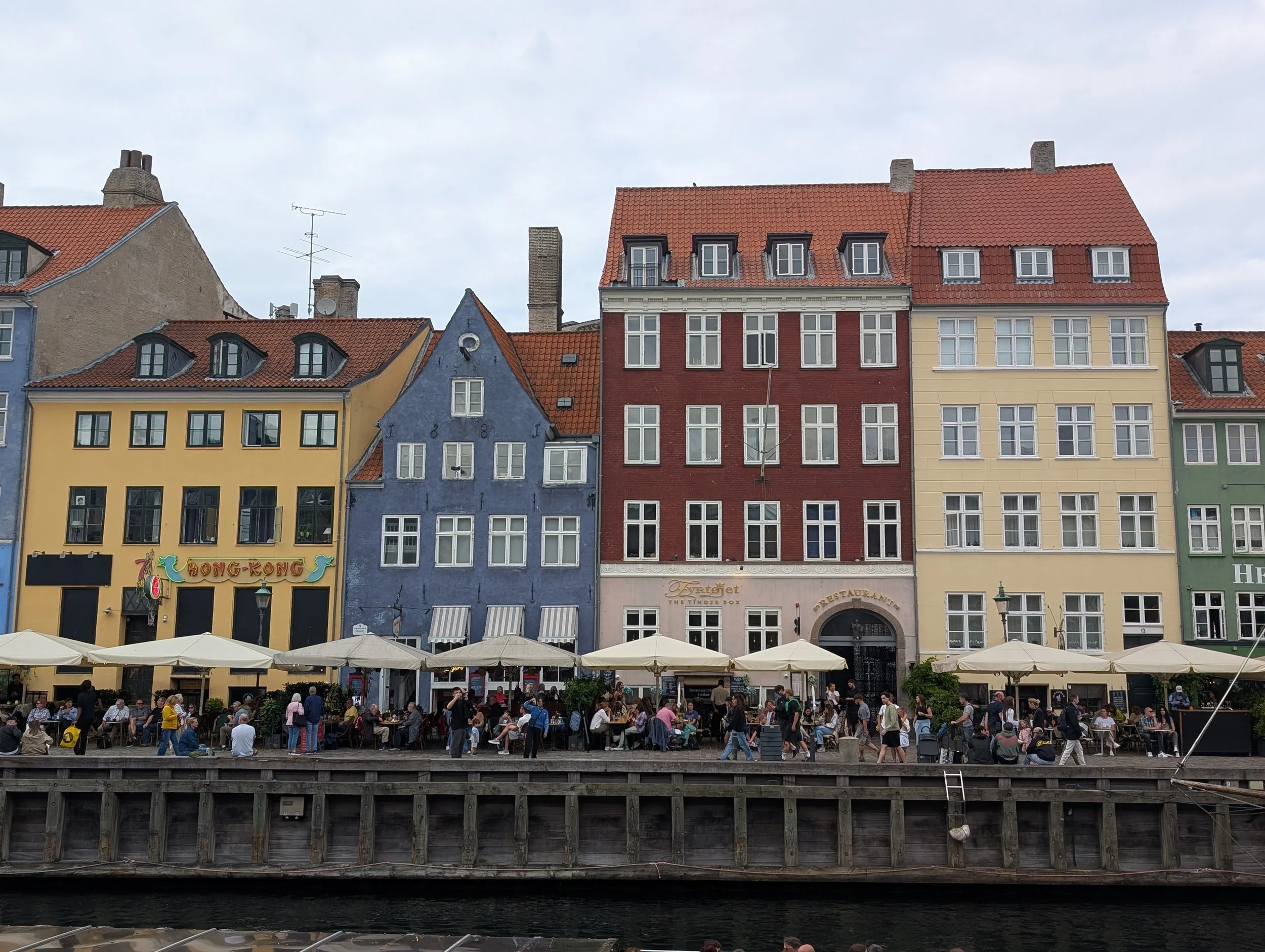People dining at outdoor cafes along a canal, with colorful European-style buildings in the background under a cloudy sky.
