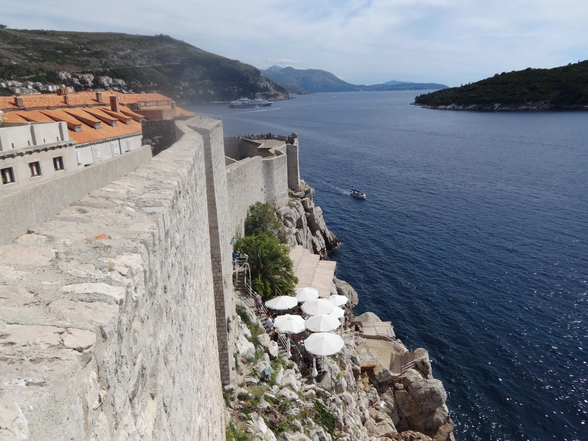 A coastal scene featuring a historical stone fortress or wall along the shoreline with a view of the sea, boats, and distant hills, and a terrace with white umbrellas and people enjoying the area.