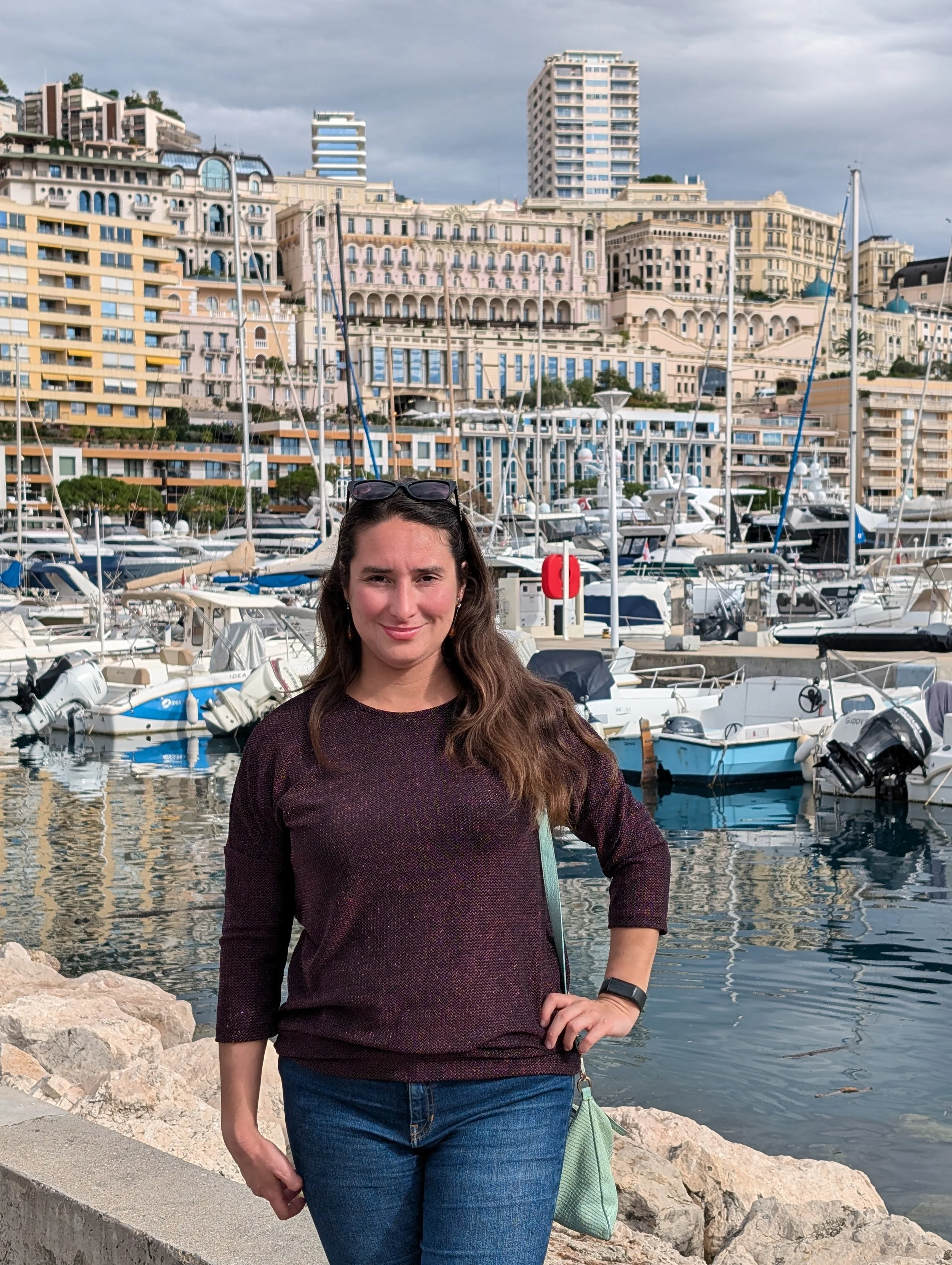 A woman with brown hair and a dark sweater standing in front of a marina filled with boats and yachts, with multi-story luxury buildings in the background, under a cloudy sky.