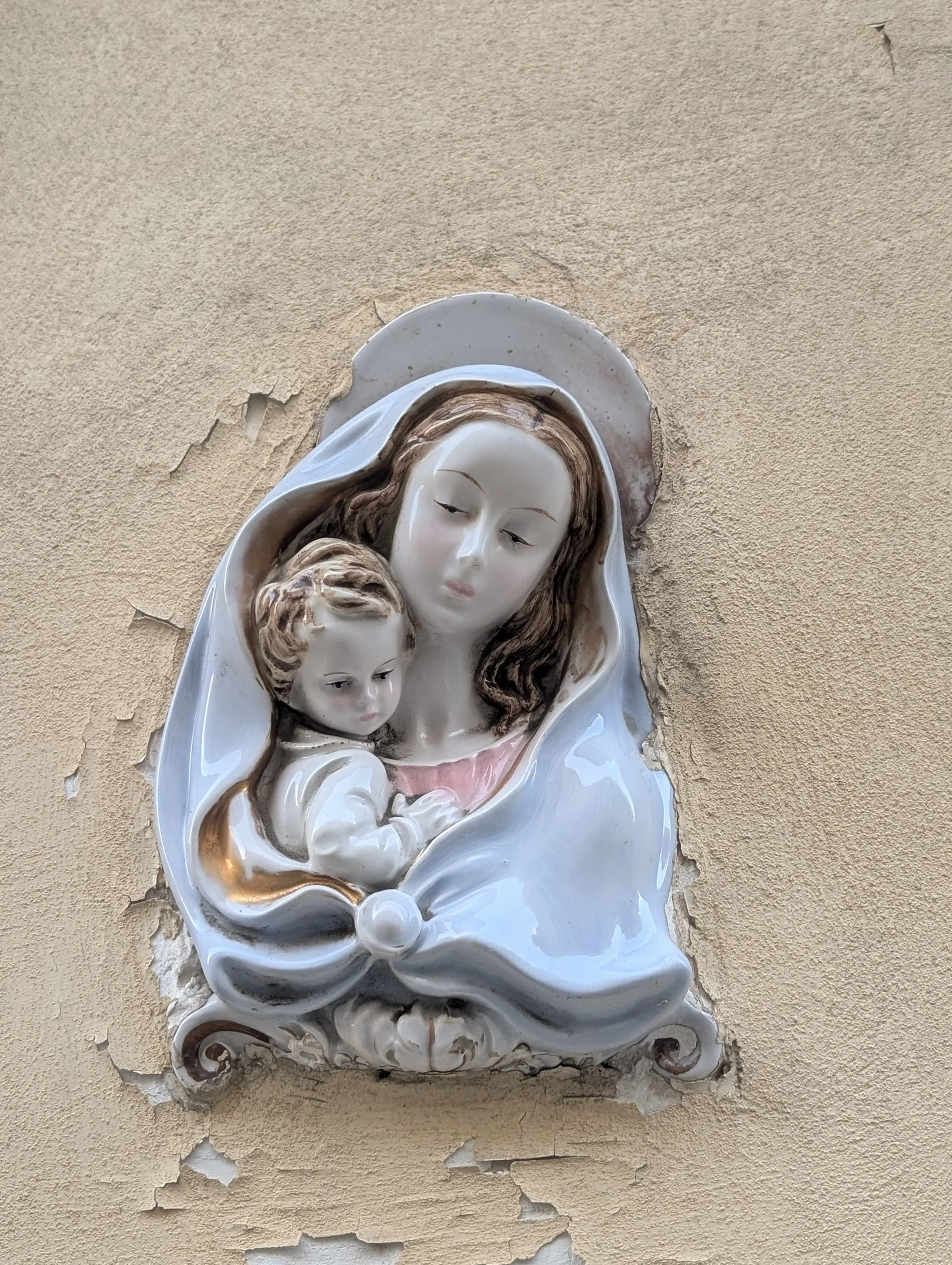 Religious wall sculpture of Mother Mary holding Baby Jesus, mounted on a chipped beige wall.