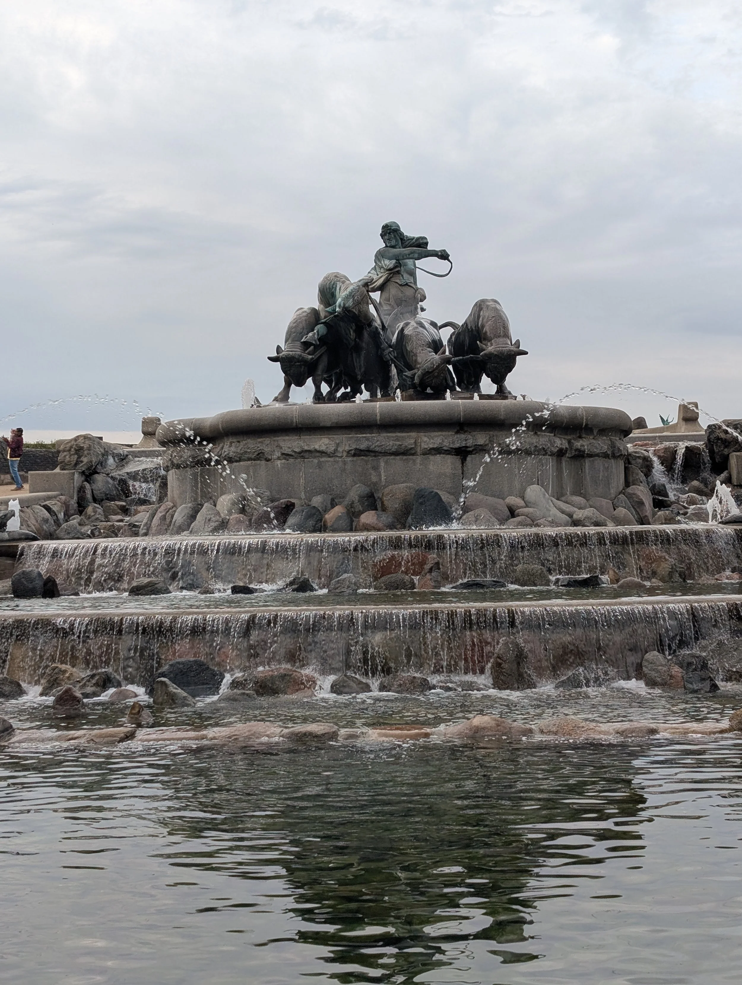 A large bronze fountain sculpture of a man riding a buffalo, surrounded by water with a cloudy sky in the background.
