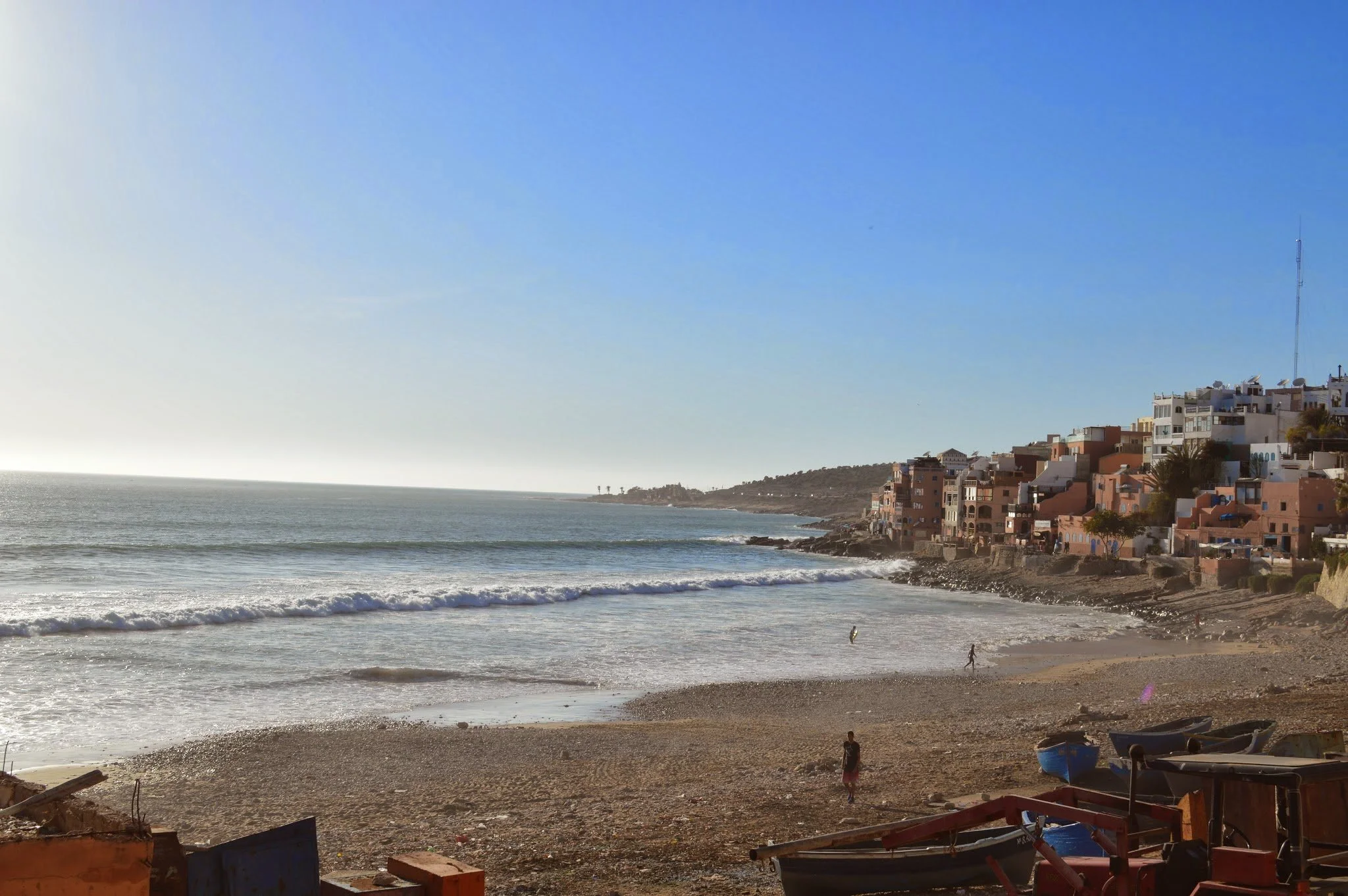 A coastal scene with a sandy beach, waves, and colourful buildings on a hillside under a clear blue sky.