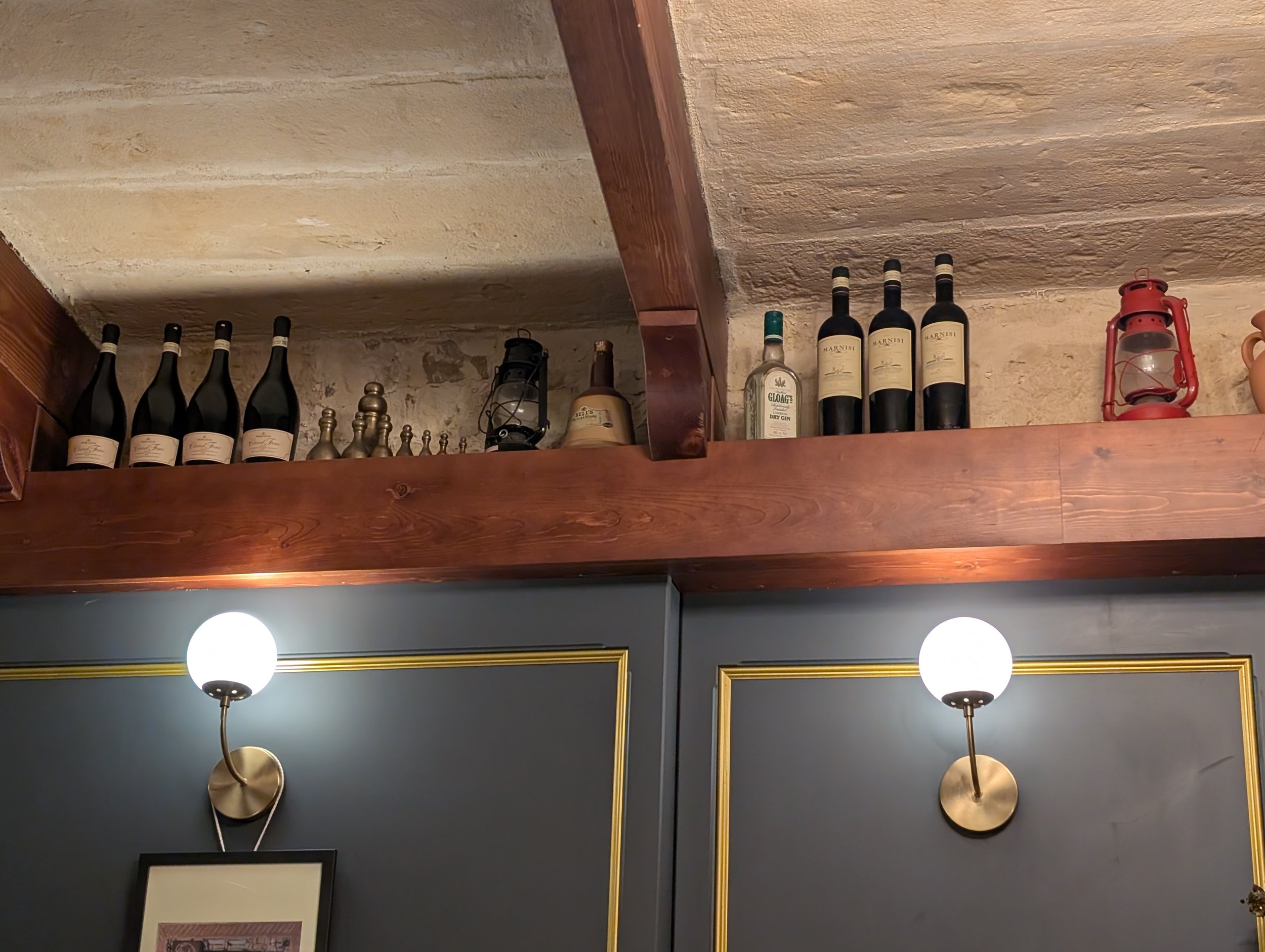 A wooden shelf attached to a ceiling with a textured stone surface, holding wine bottles, a lantern, small metallic decorative items, and a candle holder. There are lamps attached to the wall below the shelf.