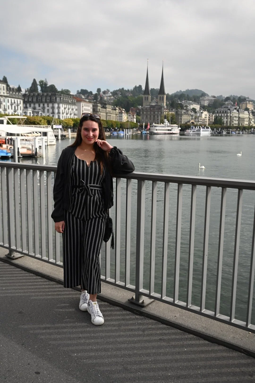 A smiling woman stands by a railing along a waterfront in a cityscape with boats, historic buildings, and two church steeples in the background.