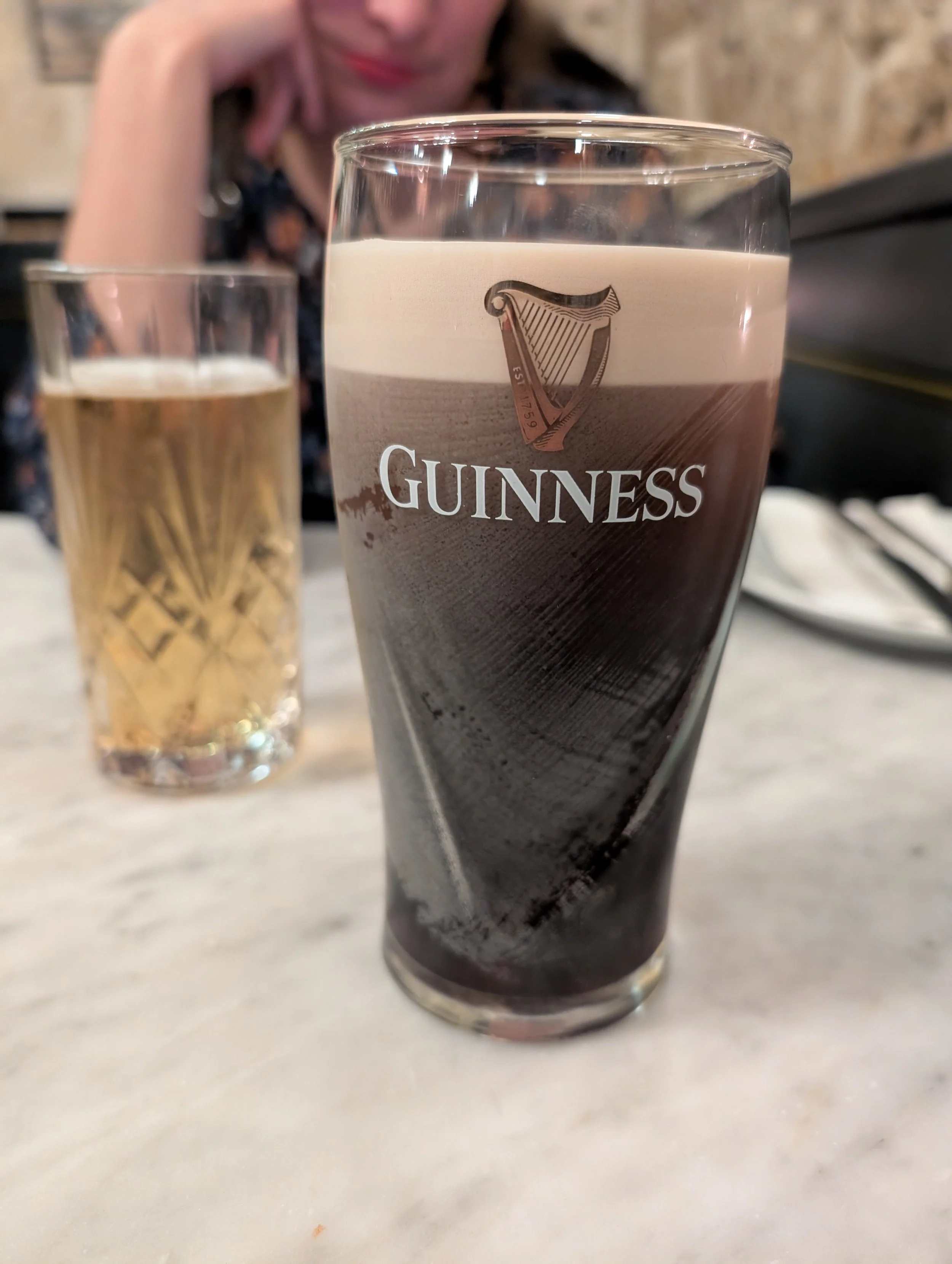 A glass of Guinness and a glass of a light-coloured beverage on a restaurant table, with a woman in the background.