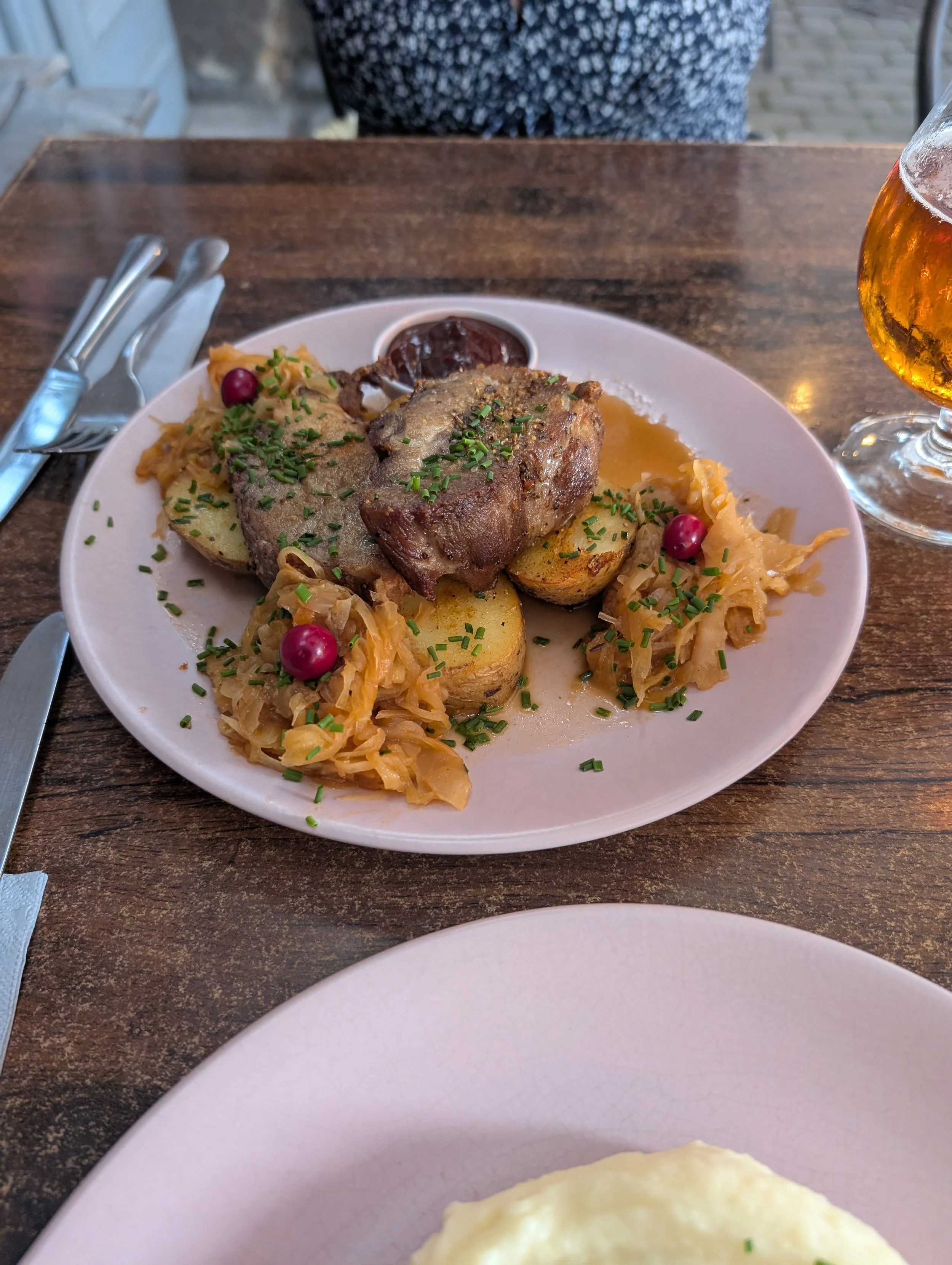 A plate of roasted meat with potatoes, sauerkraut, and cranberries served at a restaurant table, with a glass of beer and cutlery nearby.