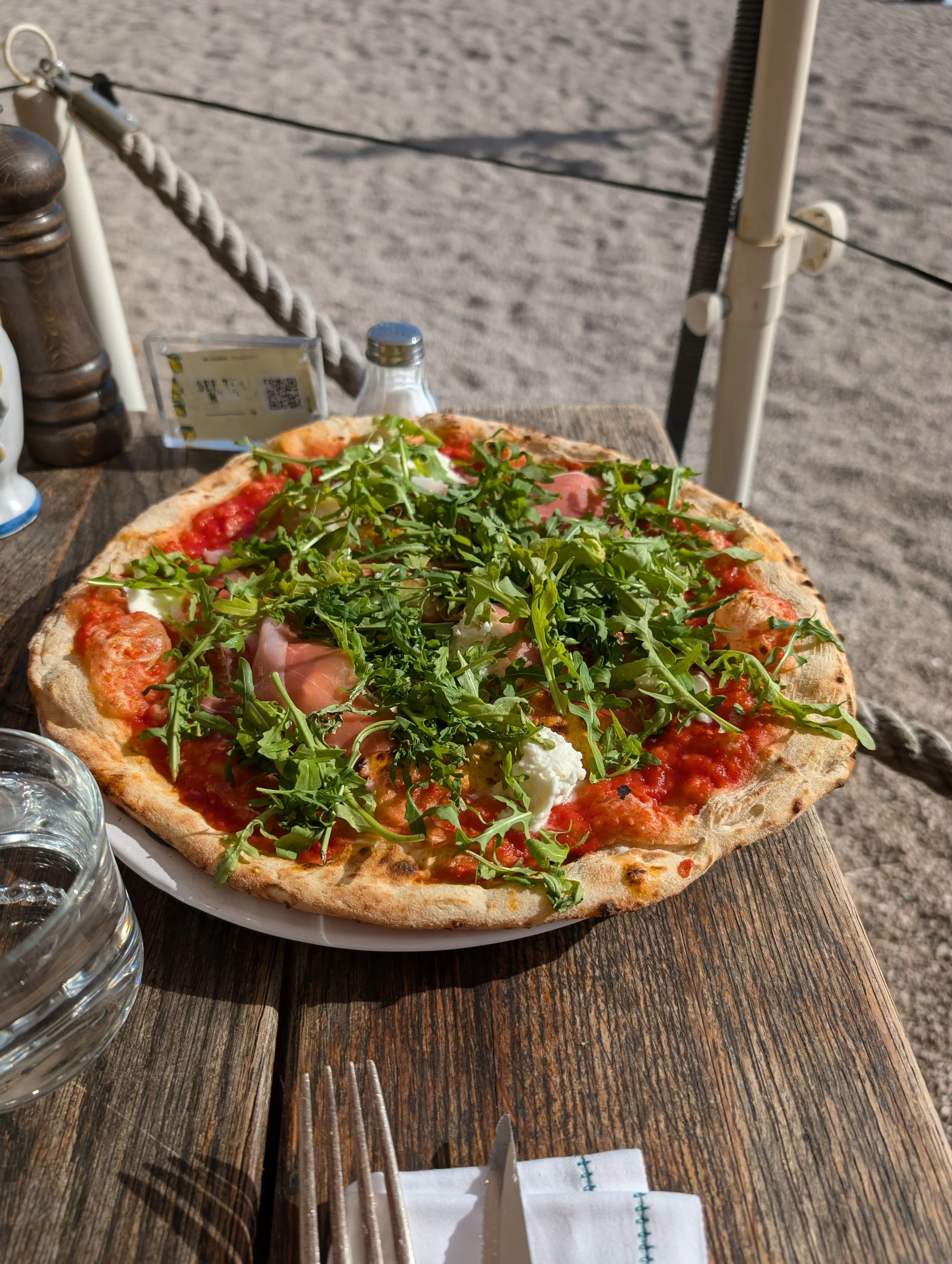 A pizza with arugula, tomatoes, and other toppings on a wooden table outdoors, with a glass of water, napkin, and utensils nearby.