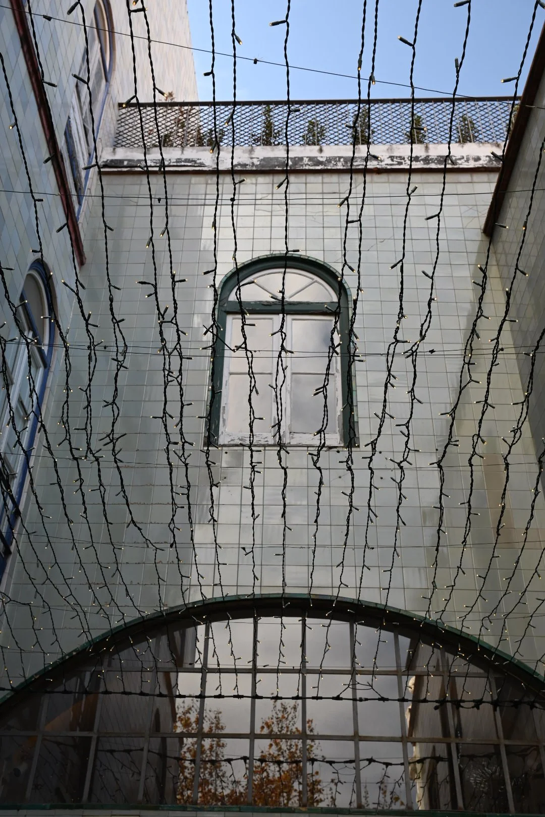 View of a building exterior with a decorative window, topped with a row of string lights hanging from above, with a small balcony or railing at the top of the image.