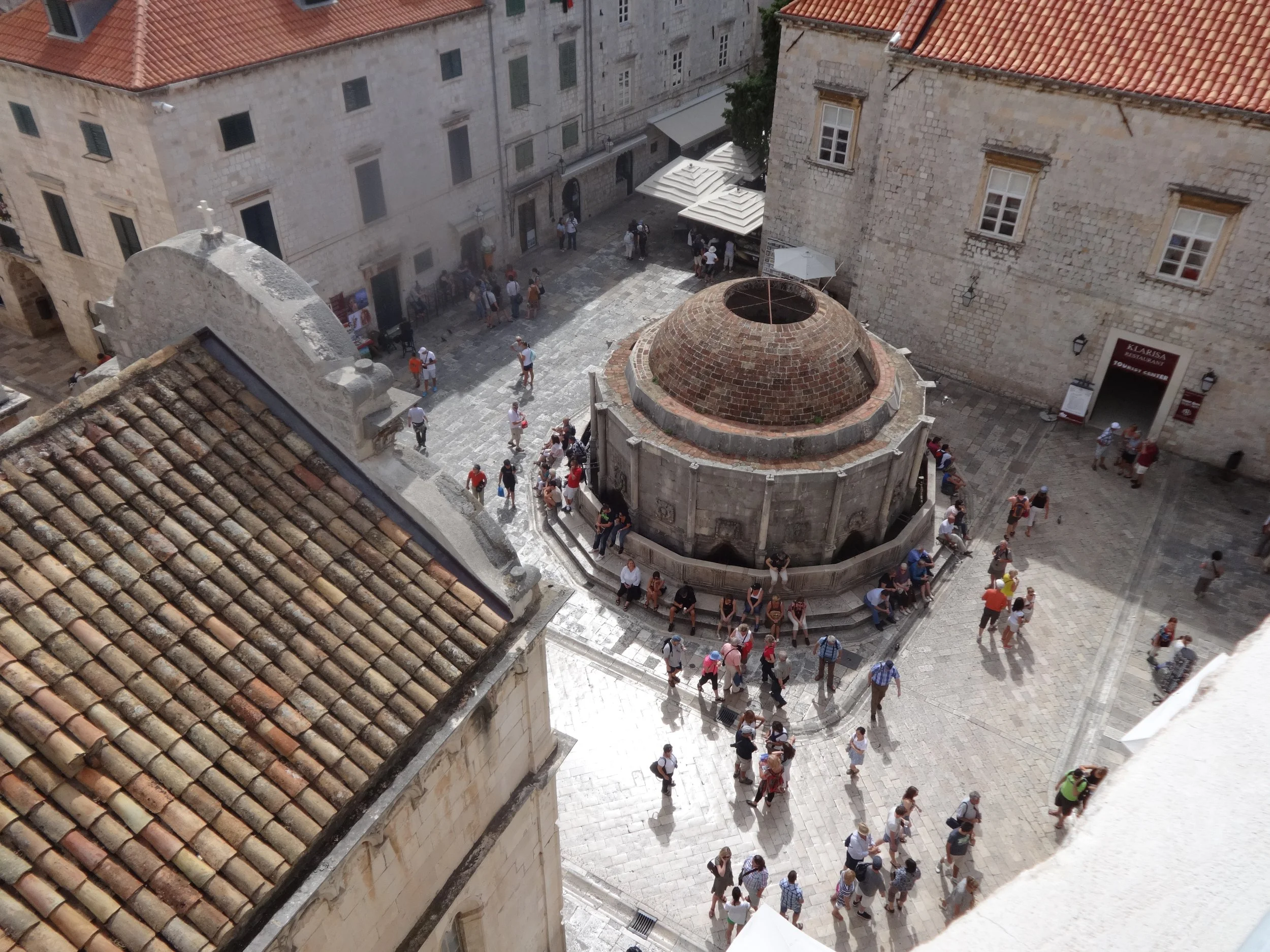 A view of a crowded cobblestone square surrounded by historic stone buildings with red-tiled roofs. People are walking and gathering in the square, and there are outdoor umbrellas shading seating areas.